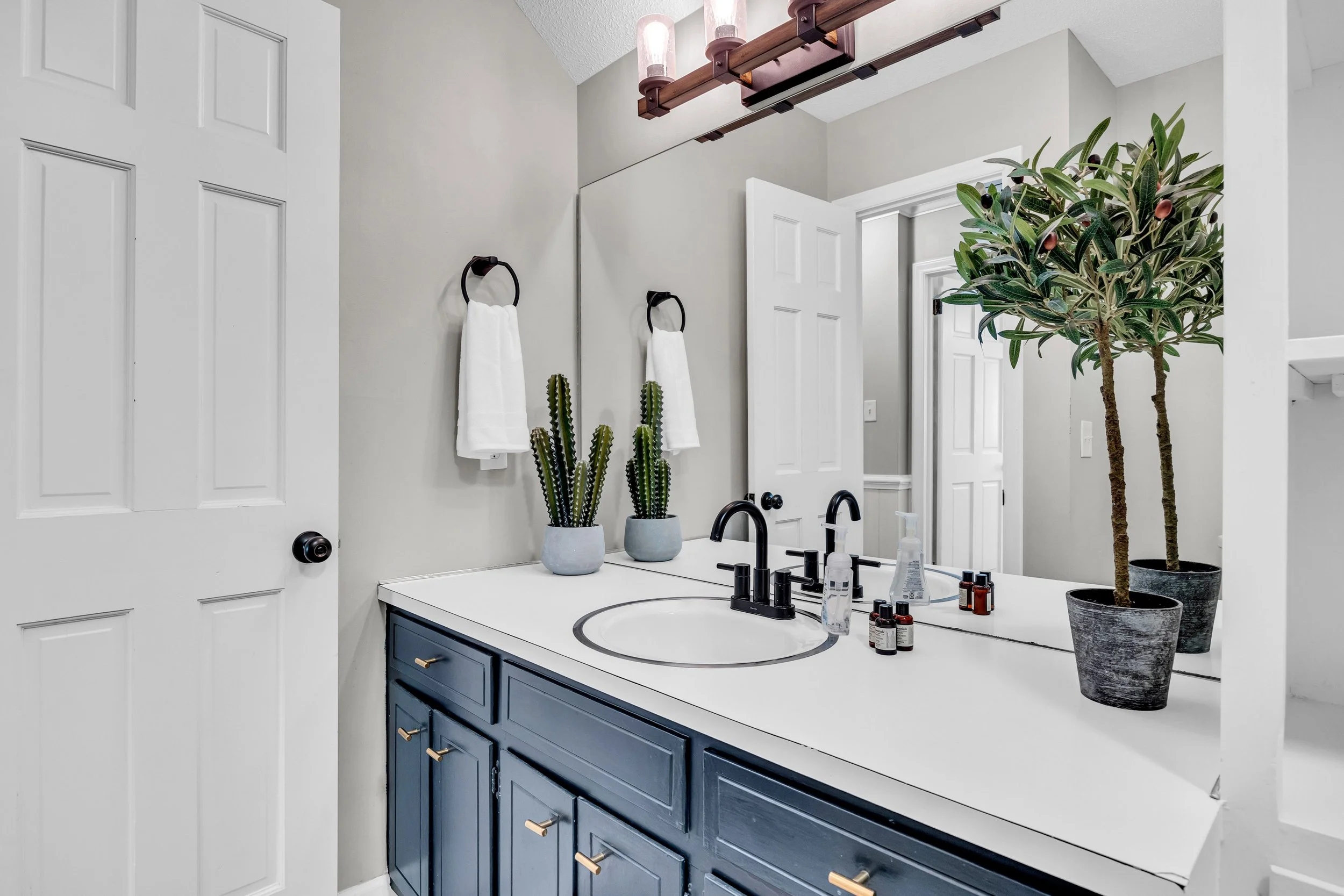 A modern bathroom vanity with a white countertop, black faucet, and navy blue cabinets with gold knobs. Decor includes potted cactus, a potted tree, and toiletry bottles. Two hand towels hang on black towel rings. There is a large mirror above the sink, with a wooden light fixture containing three bulbs above it. The walls are gray, and the bathroom has a white door and trim.