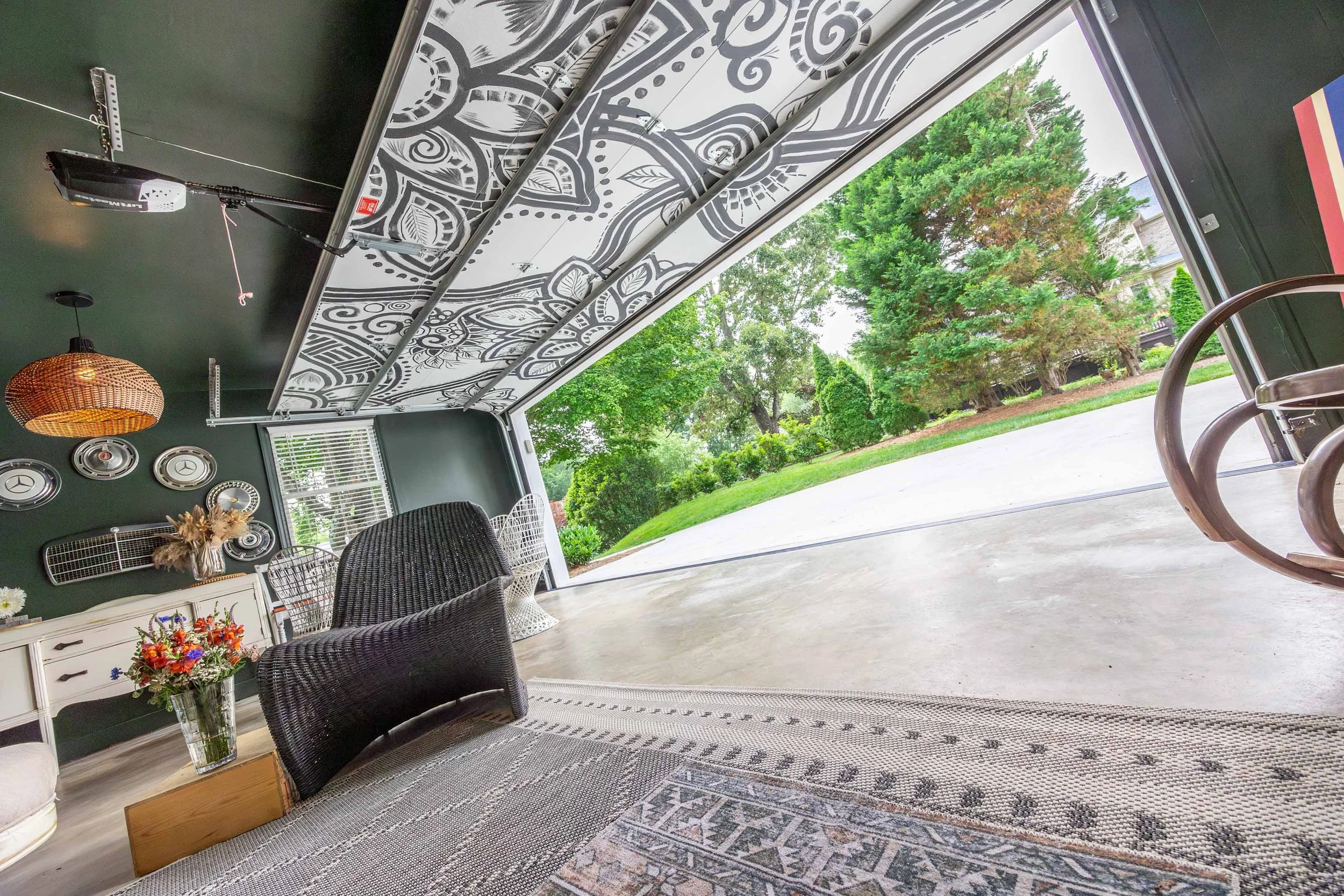 View of a living room with a large open garage door showing green trees outside. Inside, there are mixed wicker and plastic chairs, a white cabinet with floral arrangements, and various decorations on the dark green walls. The ceiling features a landscape mural and a hanging rattan light fixture.