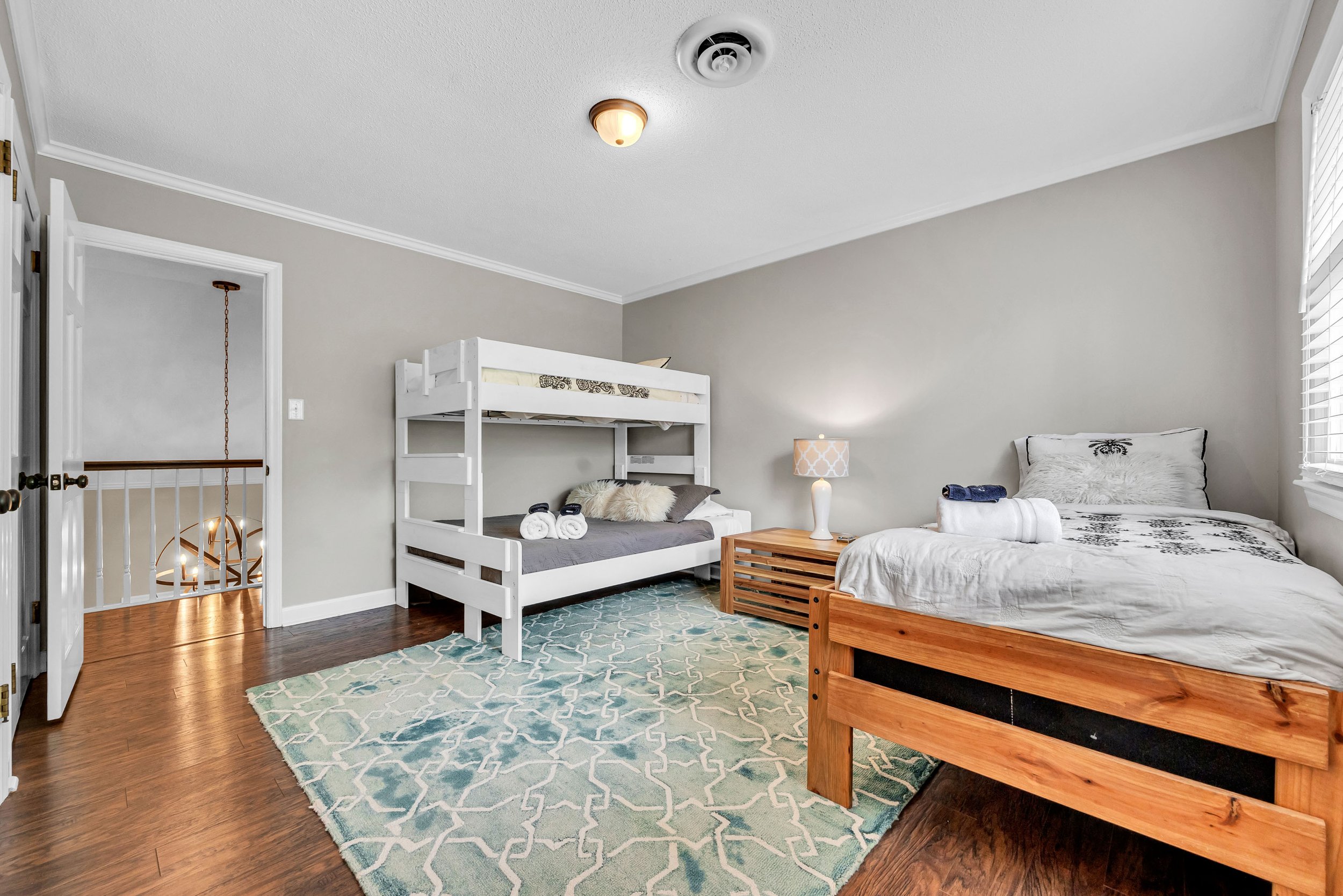 A bedroom with a bed, a bunk bed, and a wooden bed frame. The room has a green and white patterned rug, a gray wall, a table with a lamp, and rolled towels on the beds. There is a window with blinds and a ceiling light fixture.