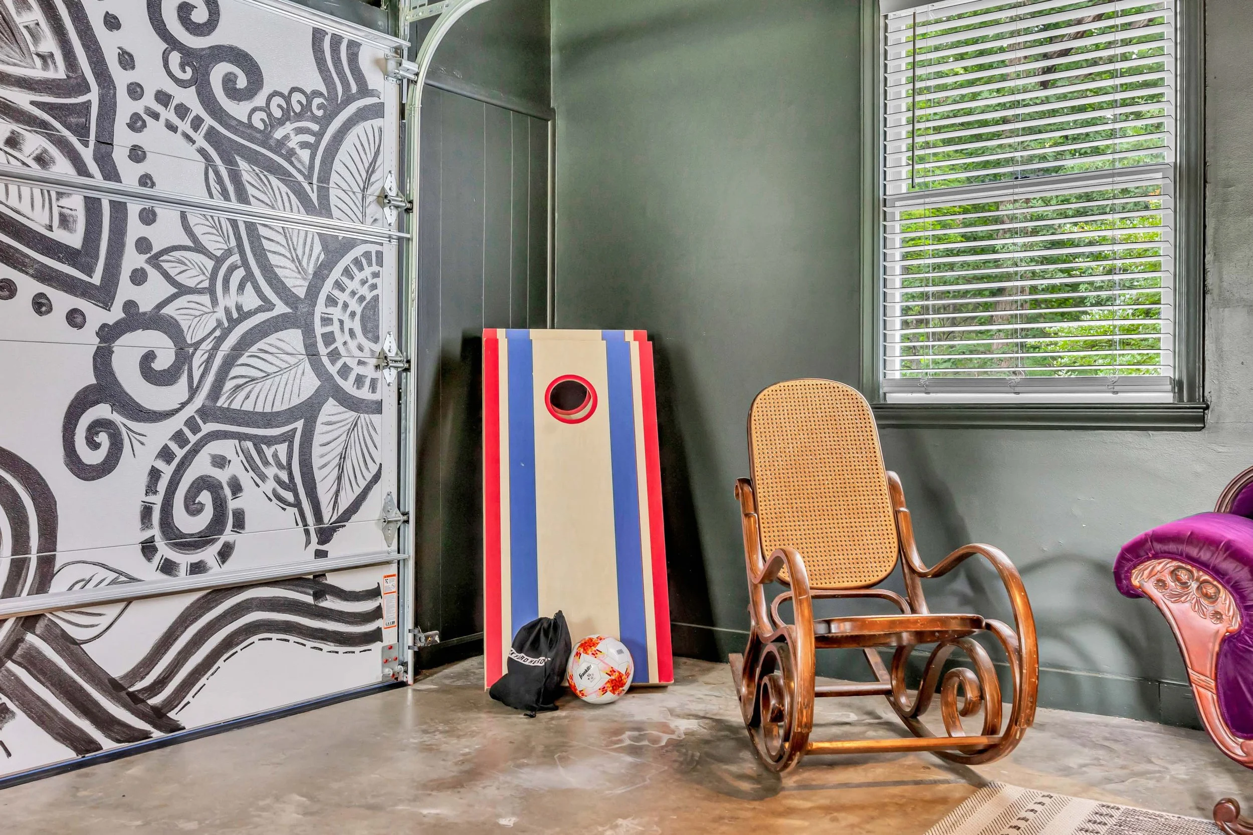 Indoor room with large window, wooden chairs, a cornhole game board, a volleyball, and miscellaneous sports equipment. The wall and floor are visible, and a garage door with black and white swirling patterns is on the left.