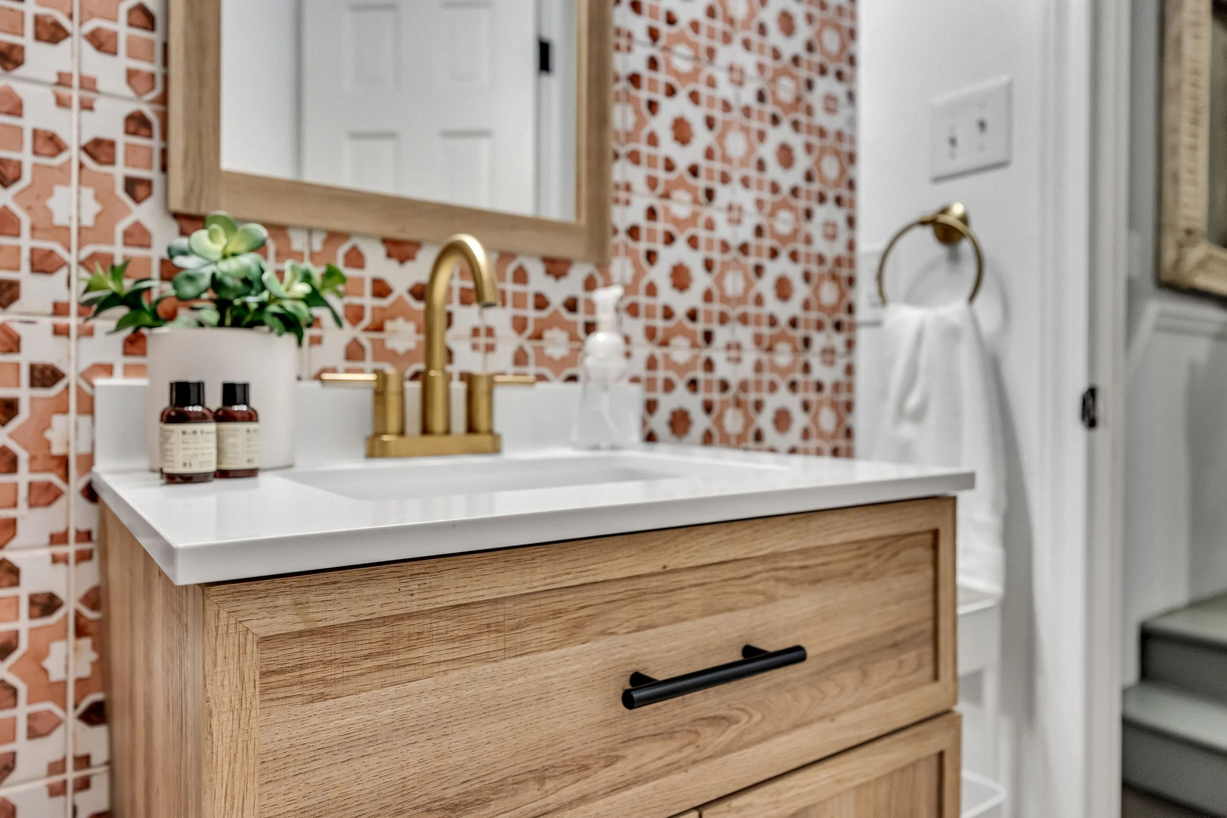 A bathroom vanity with a white countertop, a gold faucet, and a wooden cabinet below. There are two amber bottles, a small potted plant, a soap dispenser, and a glass with a wooden lid on the countertop. The background features patterned orange and white tiles, a mirror, a light switch, and a towel ring with a white towel.