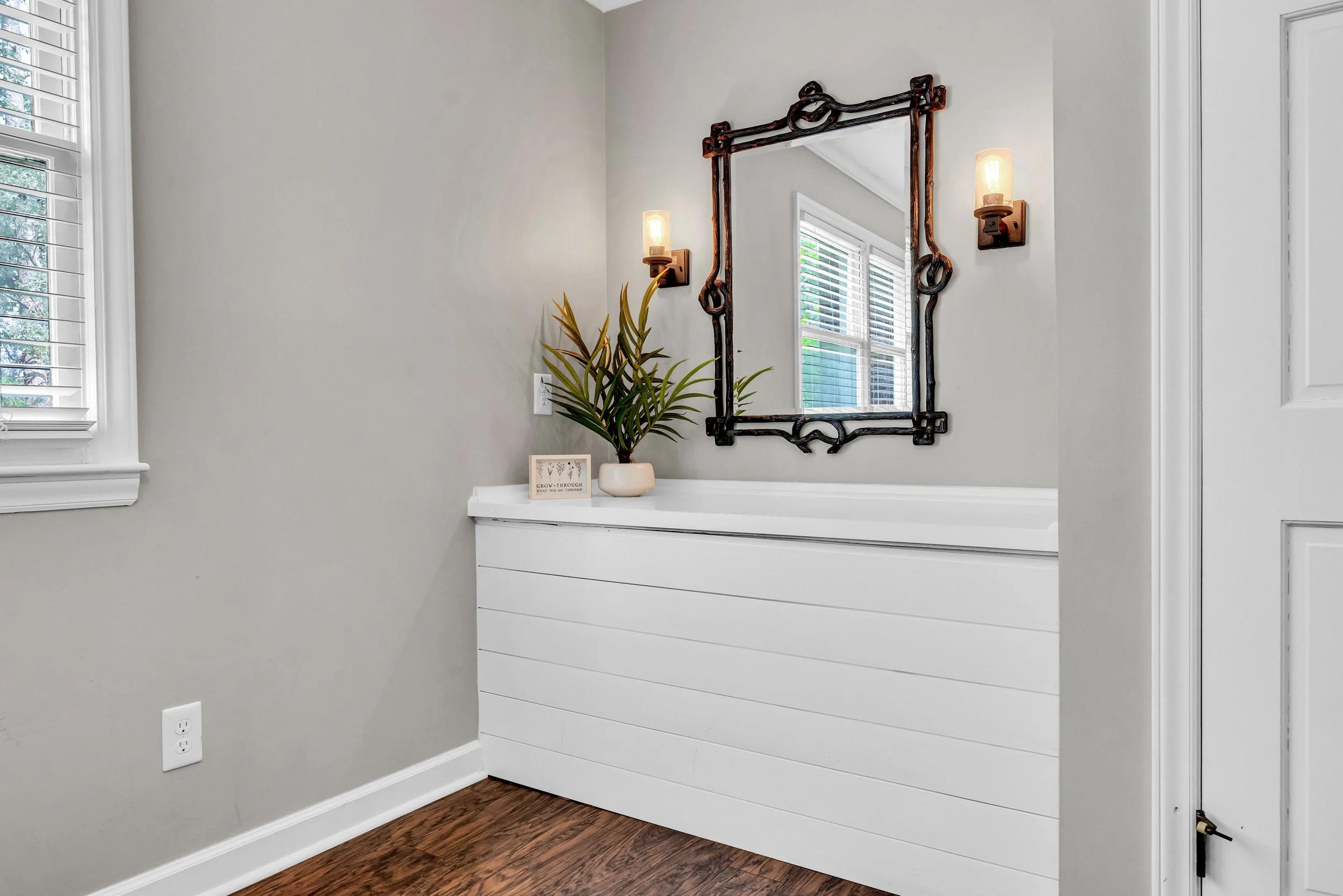 A white dresser with a potted plant on top, a picture frame, a large mirror with a decorative black frame, two wall-mounted light fixtures, a window with white blinds, and hardwood floors in a room with gray walls.