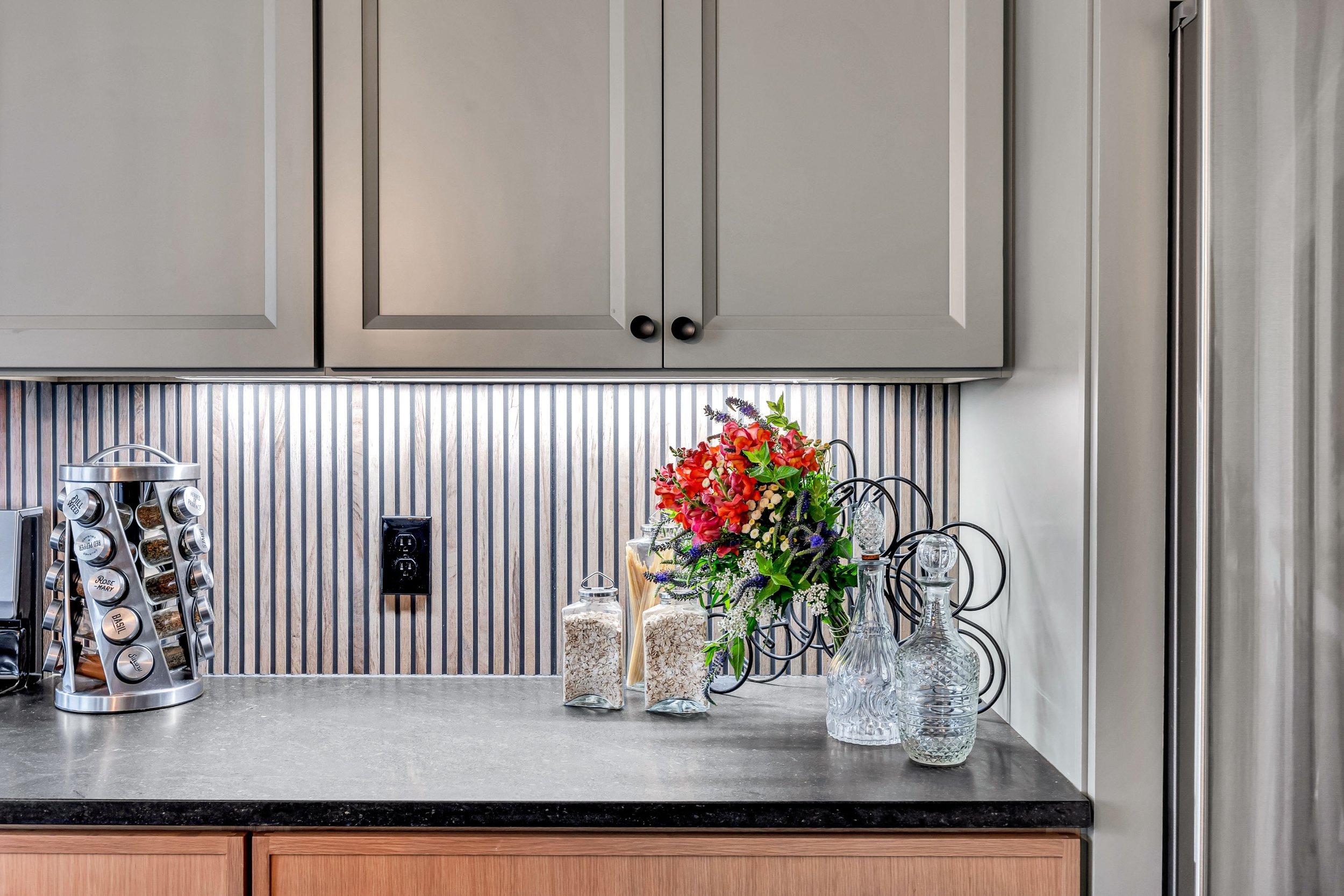 A kitchen countertop with decorative glass bottles and vases, a colorful bouquet of flowers in the center, a spice rack on the left, and gray cabinetry with black knobs above, against a striped backsplash.