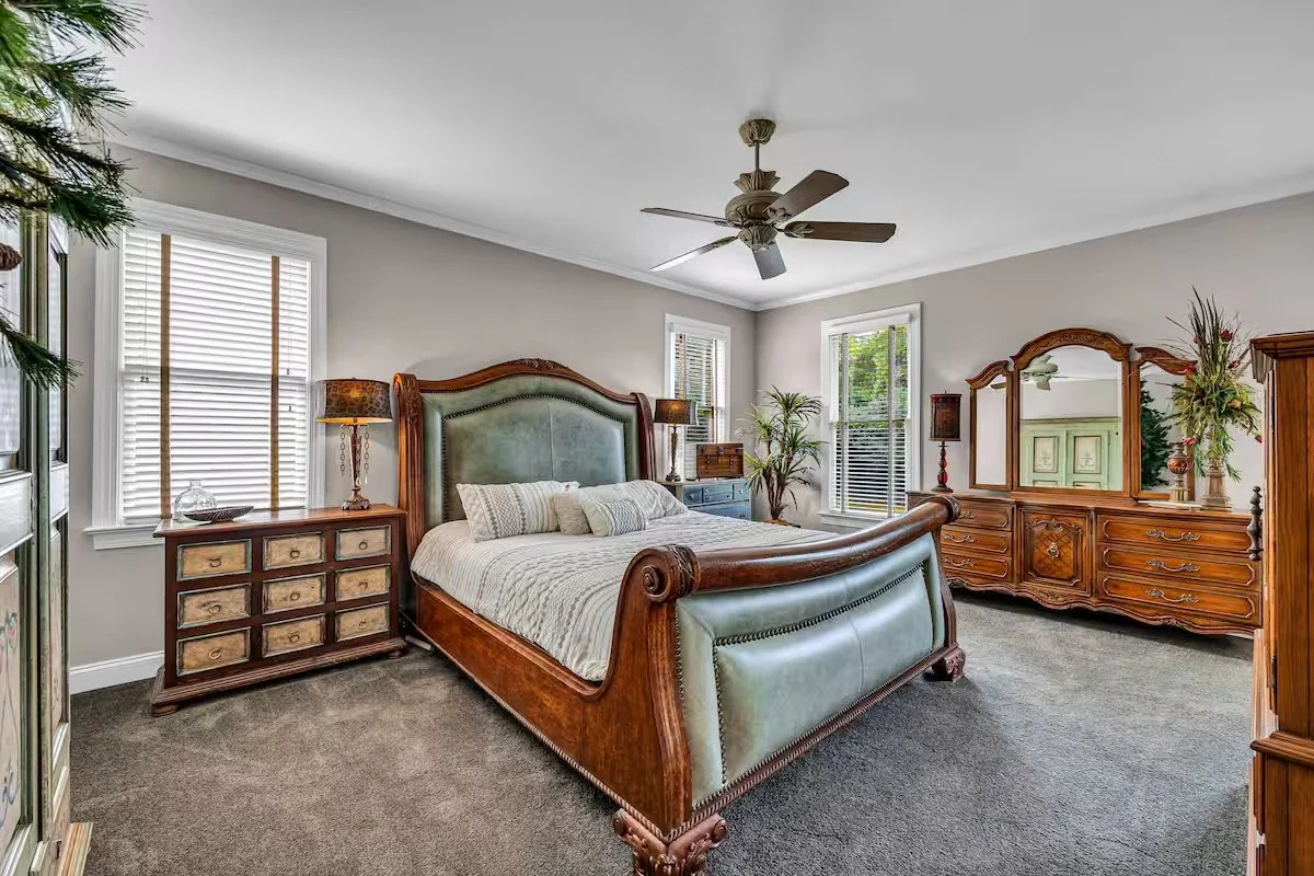 Bedroom with a large wooden bed featuring green upholstered headboard and footboard, nightstands with lamps, a wooden dresser with a mirror, and multiple windows with blinds, carpeted floor, and ceiling fan