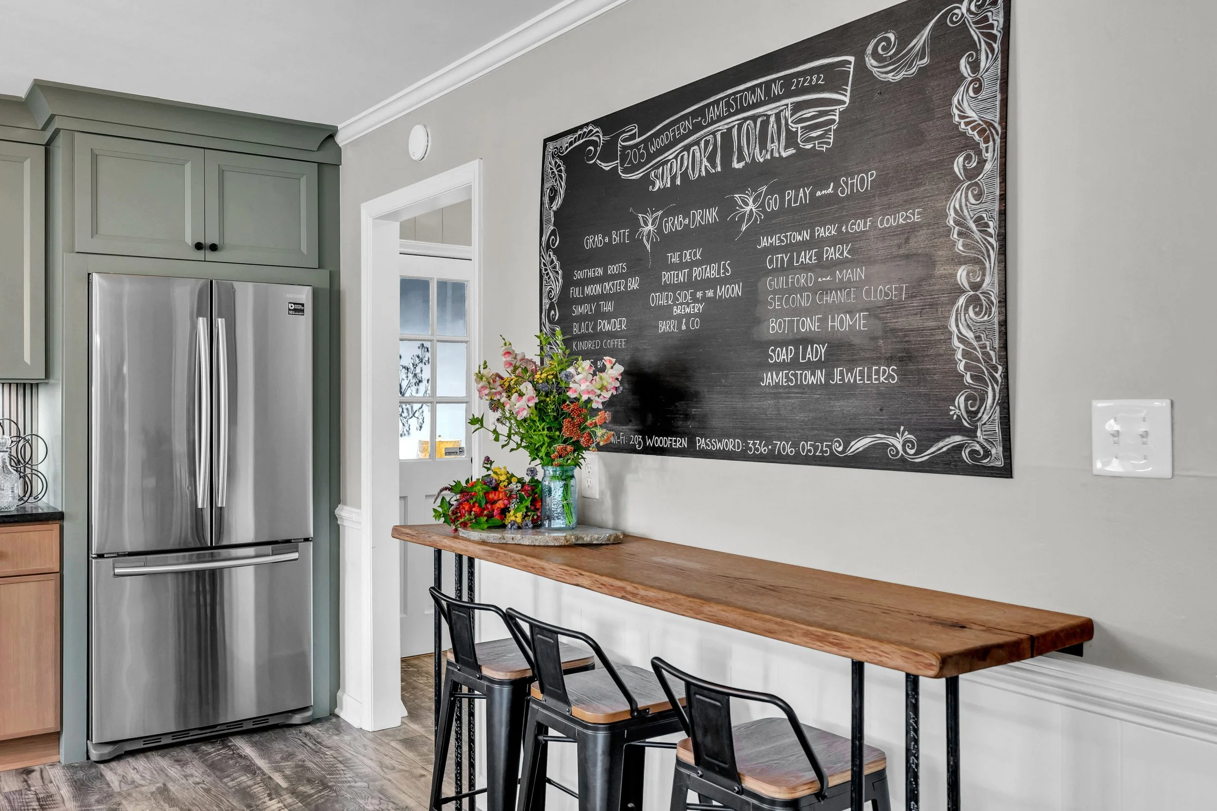 A cozy kitchen area with a large chalkboard menu on the wall listing local support businesses, a wooden table with black chairs, a bouquet of flowers, and a stainless steel refrigerator.