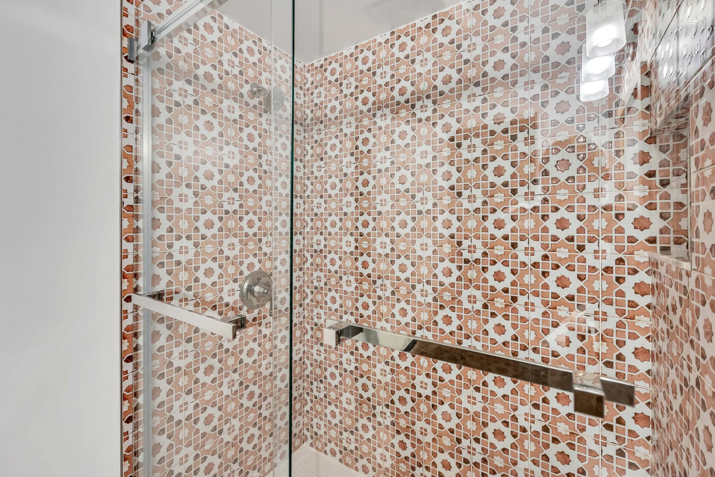 A walk-in shower with glass doors, surrounded by walls and a floor covered in patterned tiles featuring geometric shapes in shades of pink, beige, and brown.