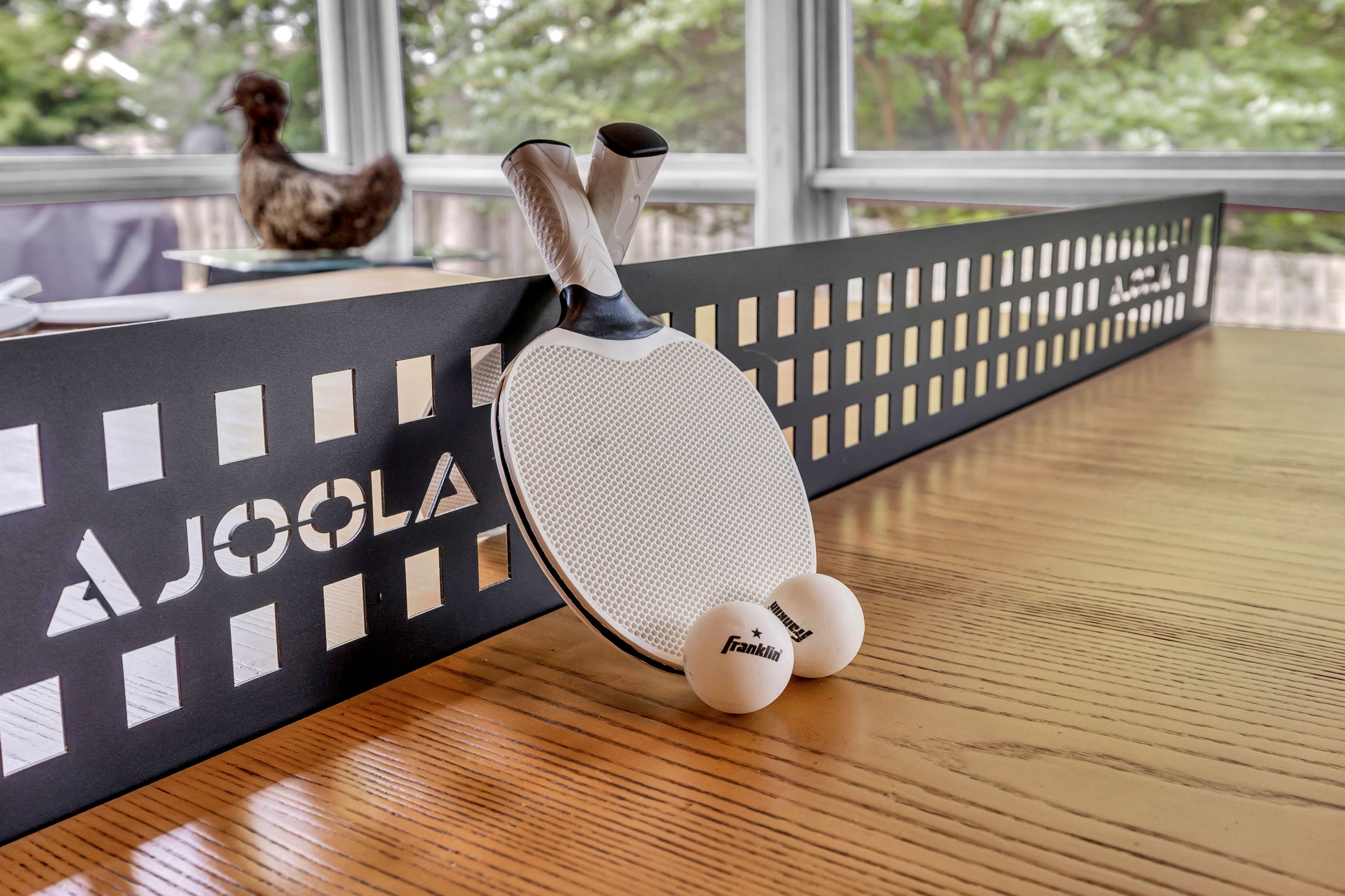 Tennis equipment including two tennis balls, a tennis racket, and a ping pong paddle placed on a wooden table near a black divider with cut-out letters, with a large window showing trees outside.