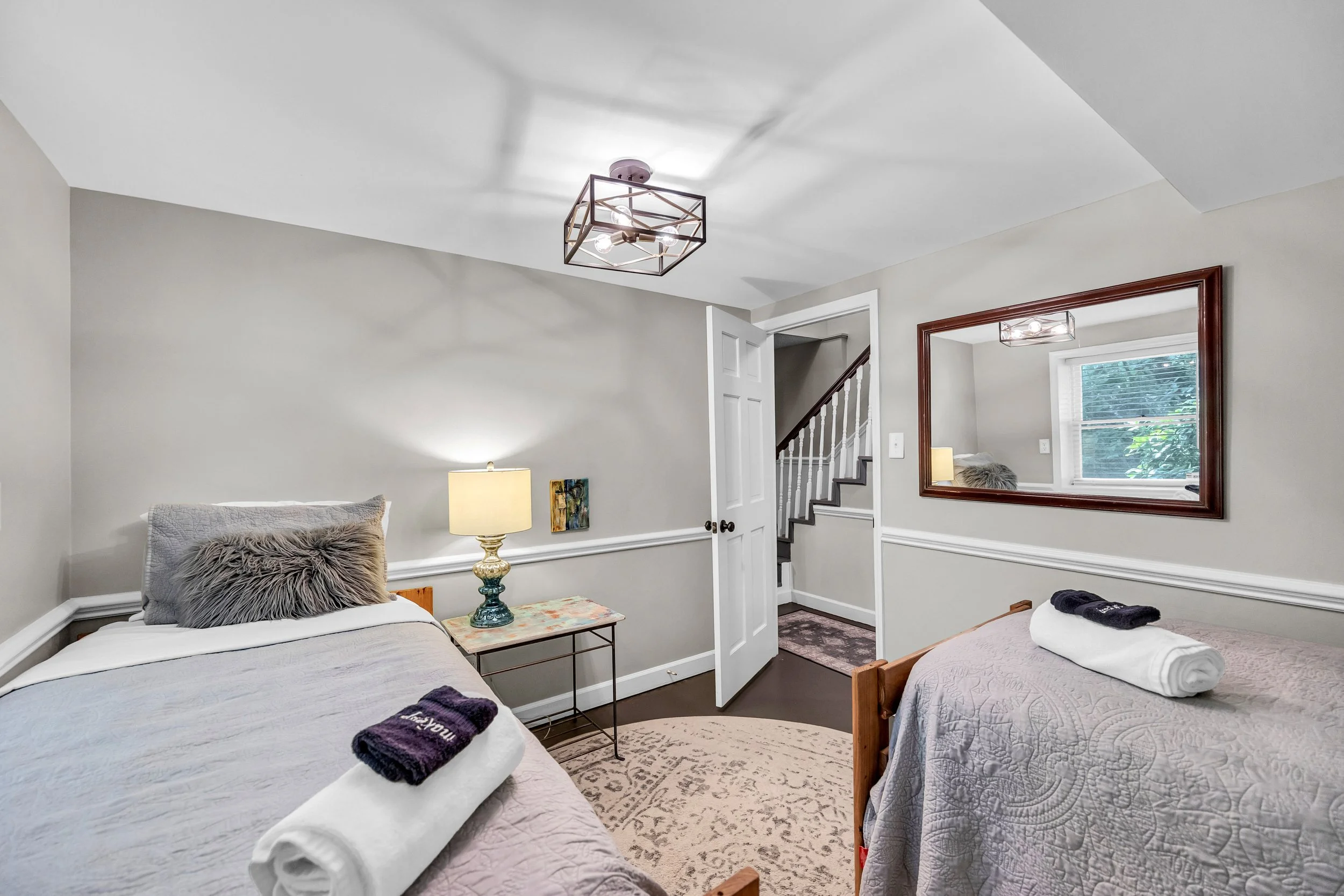 A bedroom with two twin beds, each with bed linens and rolled towels, a side table with a lamp, a large mirror on the wall, and a window with blinds. There is a staircase visible through a doorway.