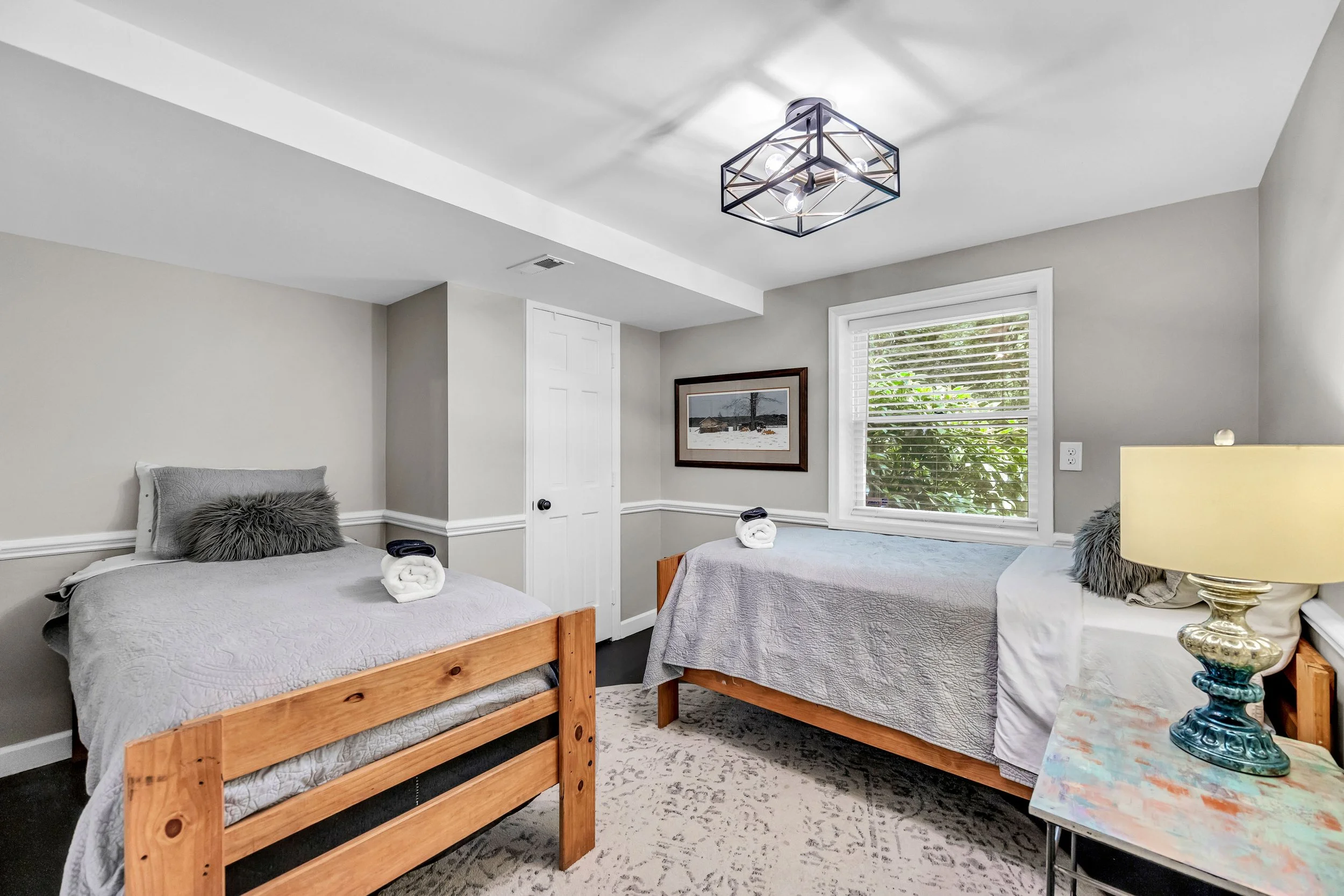 A bedroom with two beds, each with rolled towels, gray bedding, and plush gray pillows. There is a window bringing in natural light, a framed picture on the wall, a door, and a nightstand with a colorful lamp.