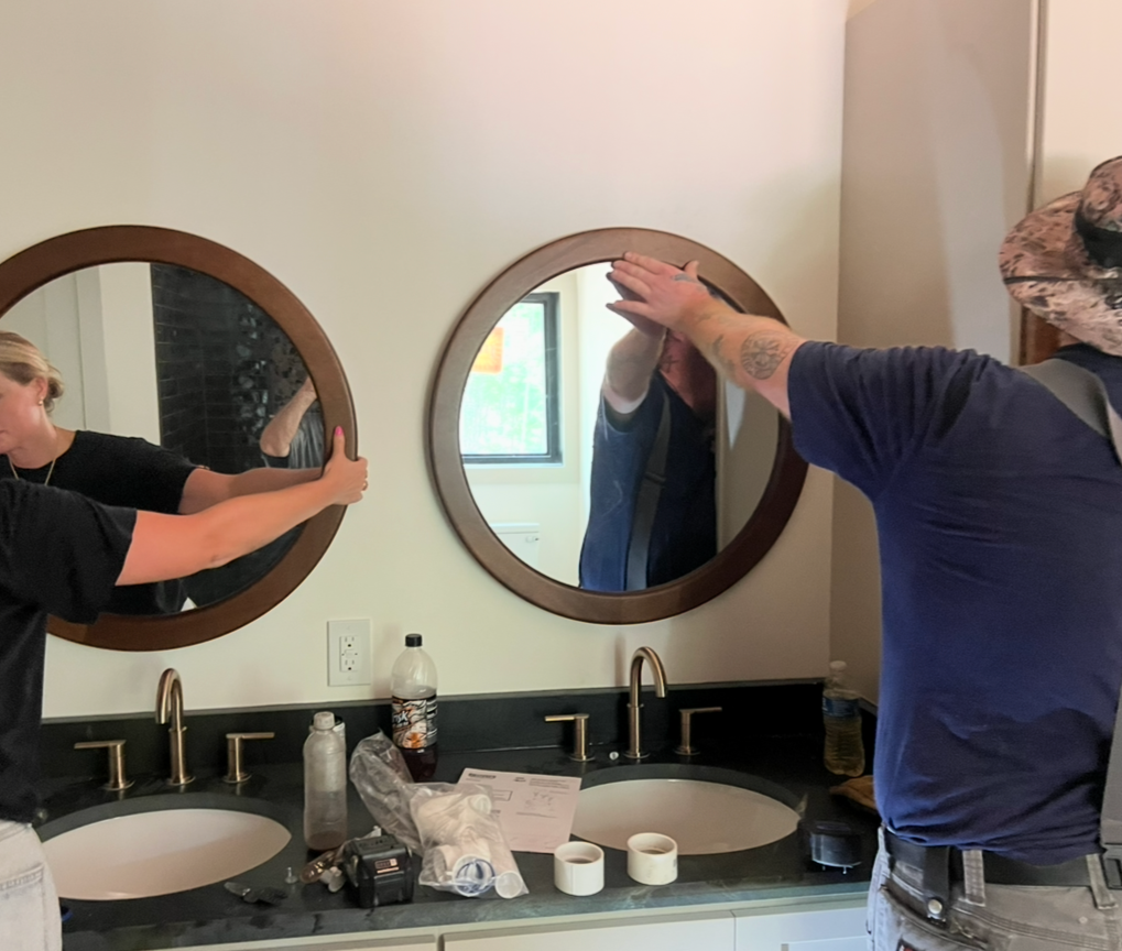 Two people wearing face masks and hats are preparing to remove or adjust a mirror above a bathroom sink. They are holding the wooden frame of the mirror, which is reflecting a window and a person in the background.