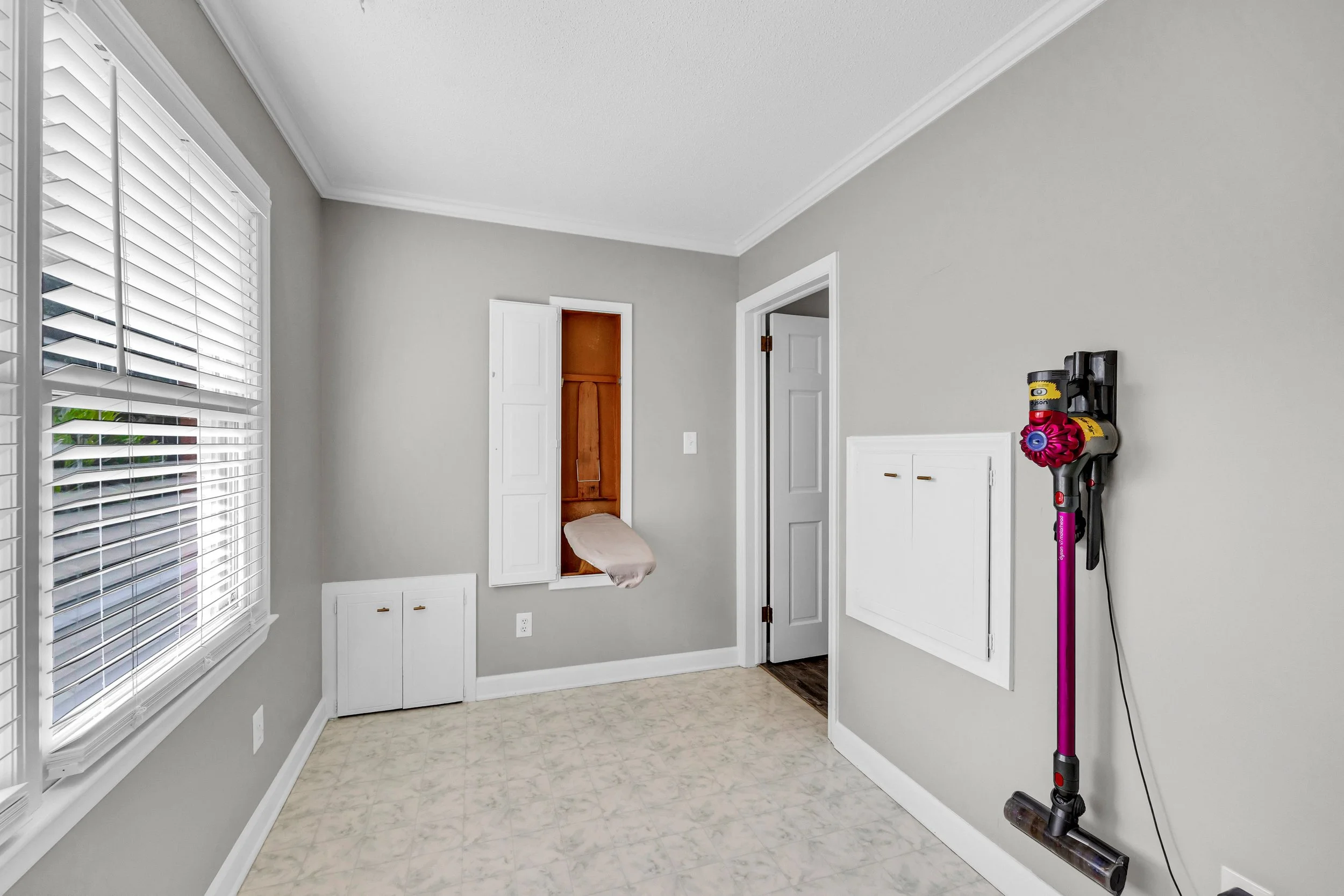 A room with a window with white blinds, a closet with double white doors, a small cabinet below the closet, and a Dyson vacuum cleaner mounted on the wall.