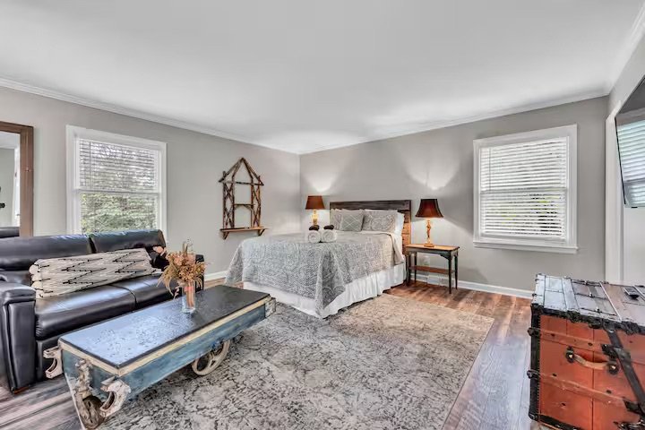 A spacious bedroom with a large bed, two nightstands with lamps, a black leather sofa, a coffee table with flowers, windows with blinds, a textured rug, and a vintage trunk in the corner.