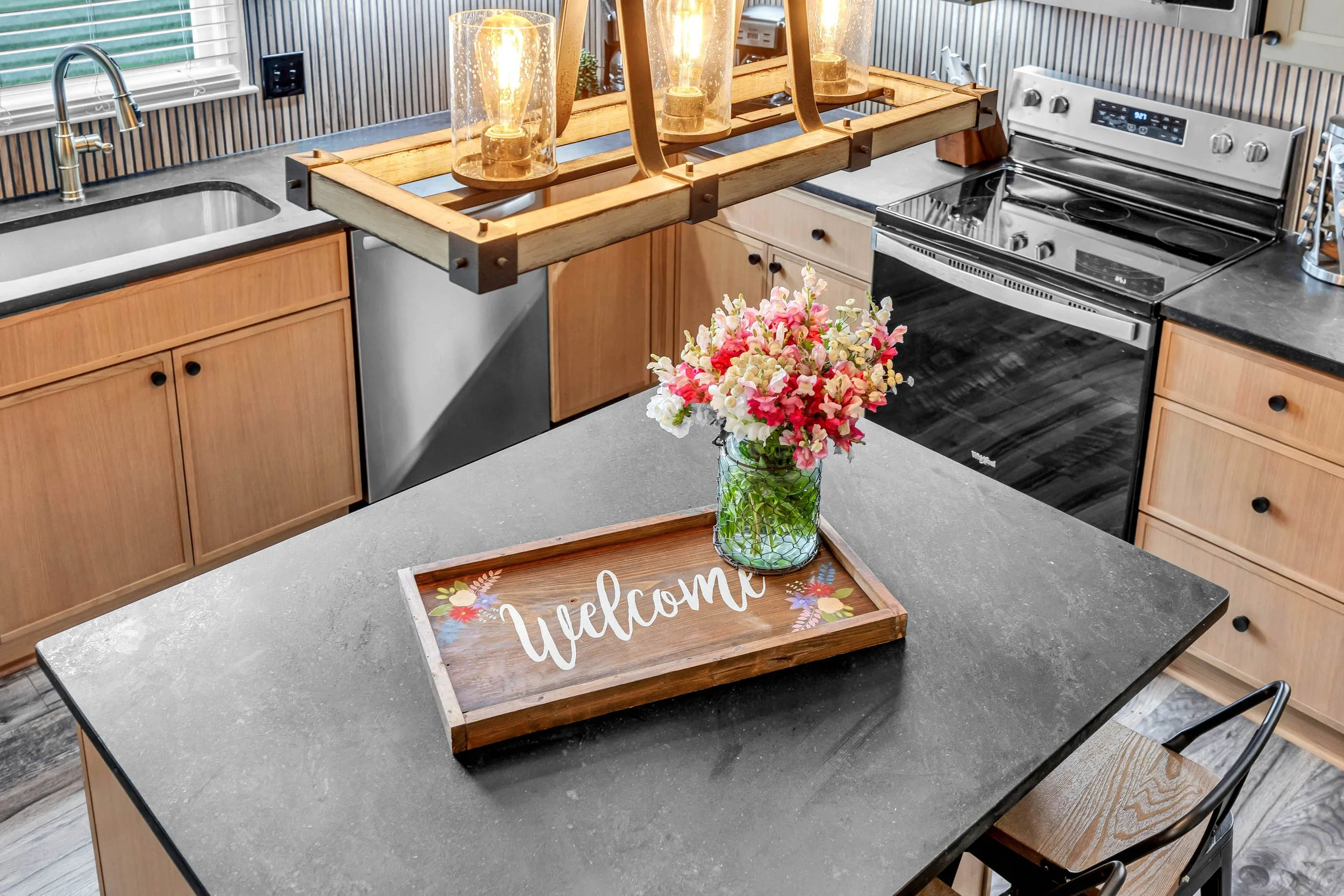 A kitchen counter with a decorative tray, a vase of pink and white flowers, and a wooden 'Welcome' sign with floral designs.