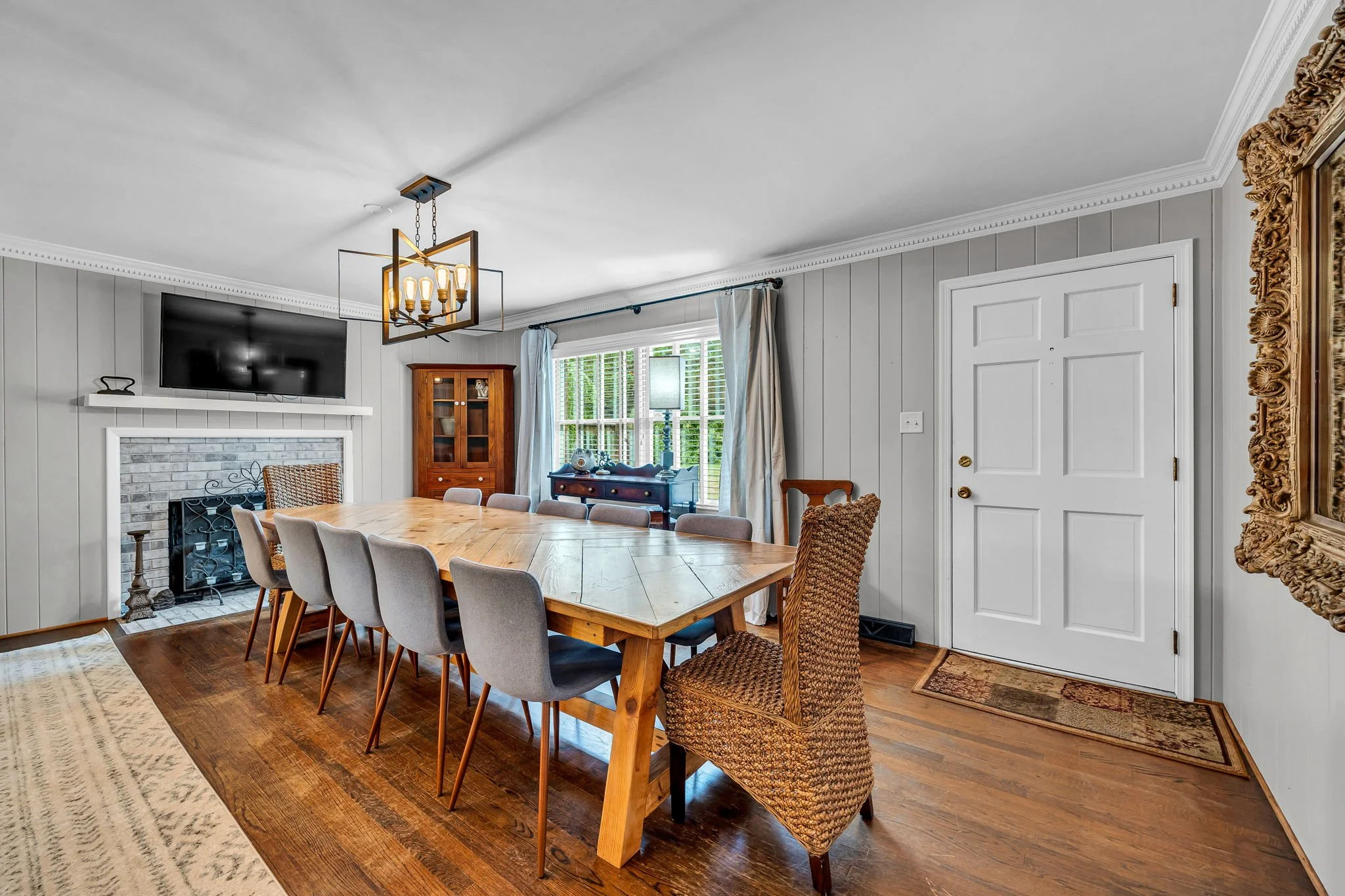 A dining room with a wooden table surrounded by beige chairs. There is a fireplace, a mounted flat-screen TV, and a large mirror with an ornate gold frame. A window with white curtains lets in natural light.