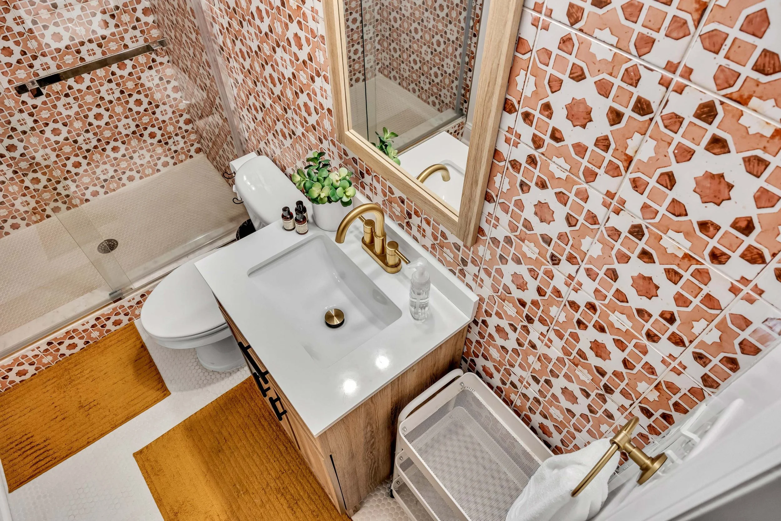 A bathroom with red and white patterned tiles on the walls, a white sink with a gold faucet, a mirror framed in wood, a small shower with a glass door, a white toilet, and a drying rack with laundry. There are small bottles on the sink, a potted plant, a brown bath mat, and a yellow rug.