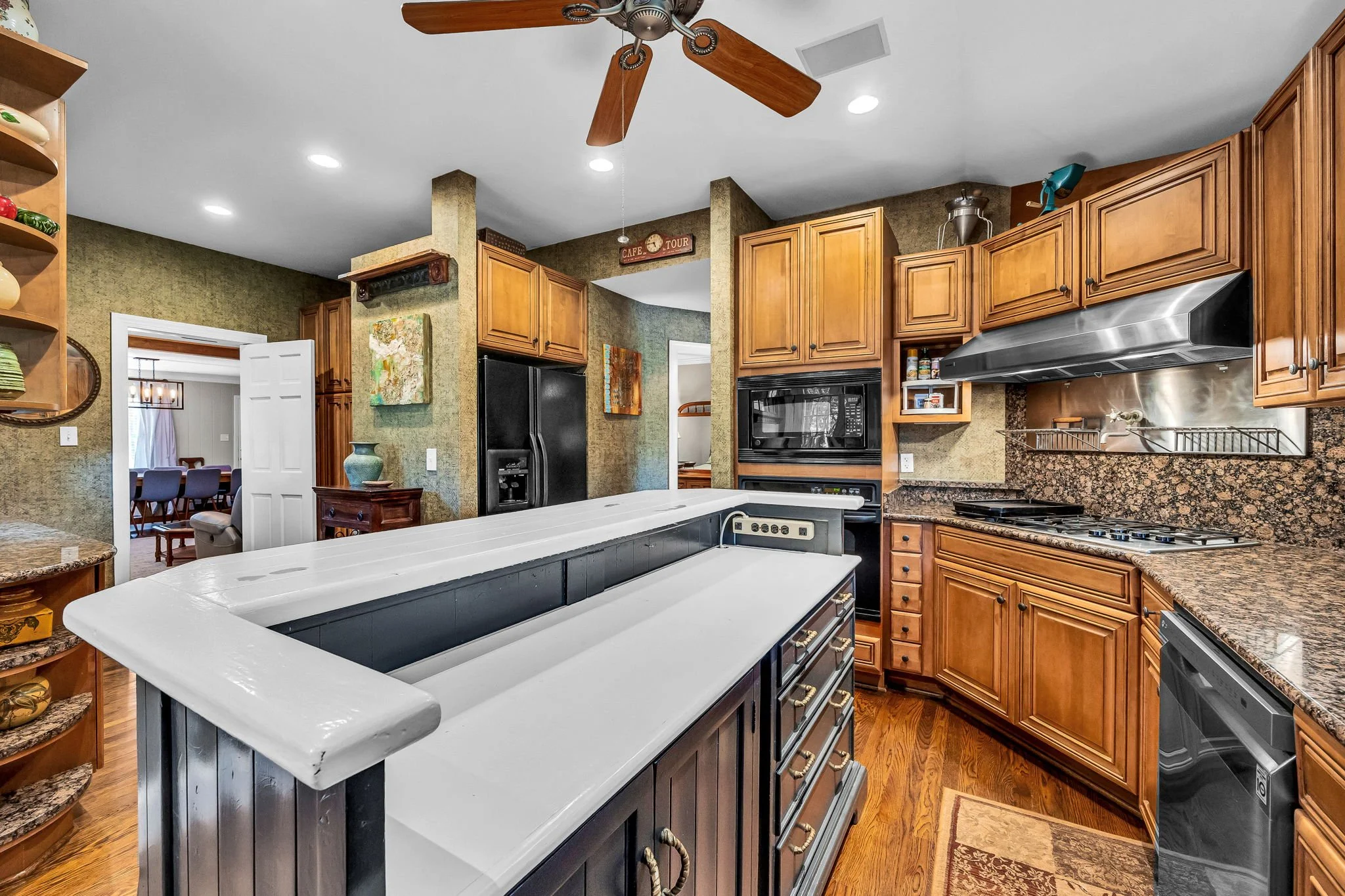 A kitchen with wooden cabinets, a granite countertop, a black microwave oven, a black refrigerator, and a white island with a stove. There is a ceiling fan with wooden blades, and a doorway leading to a dining room.
