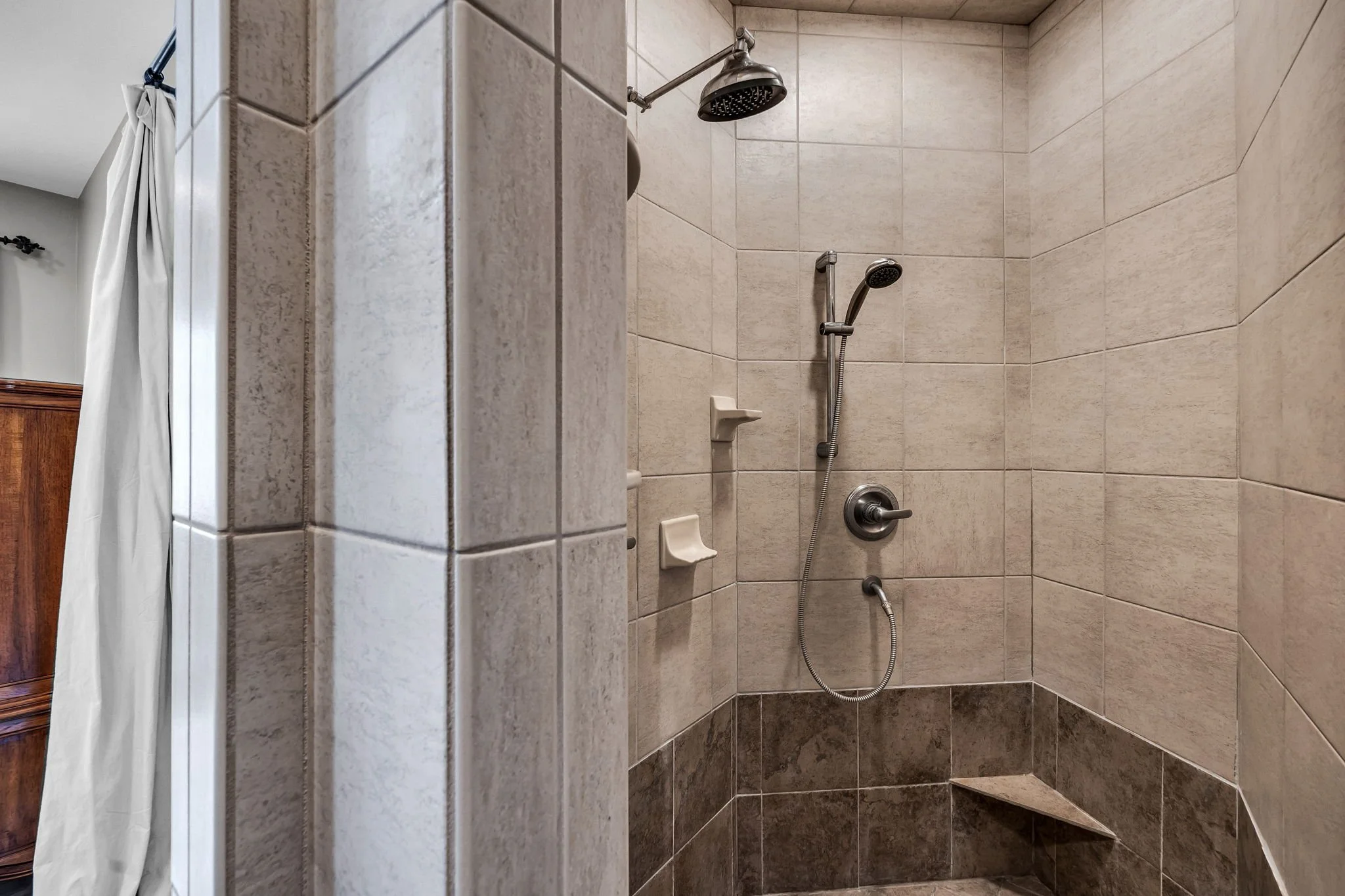 Tile shower stall with wall-mounted showerhead, handheld shower attachment, built-in soap holders, and a tiled bench with a small built-in shelf.