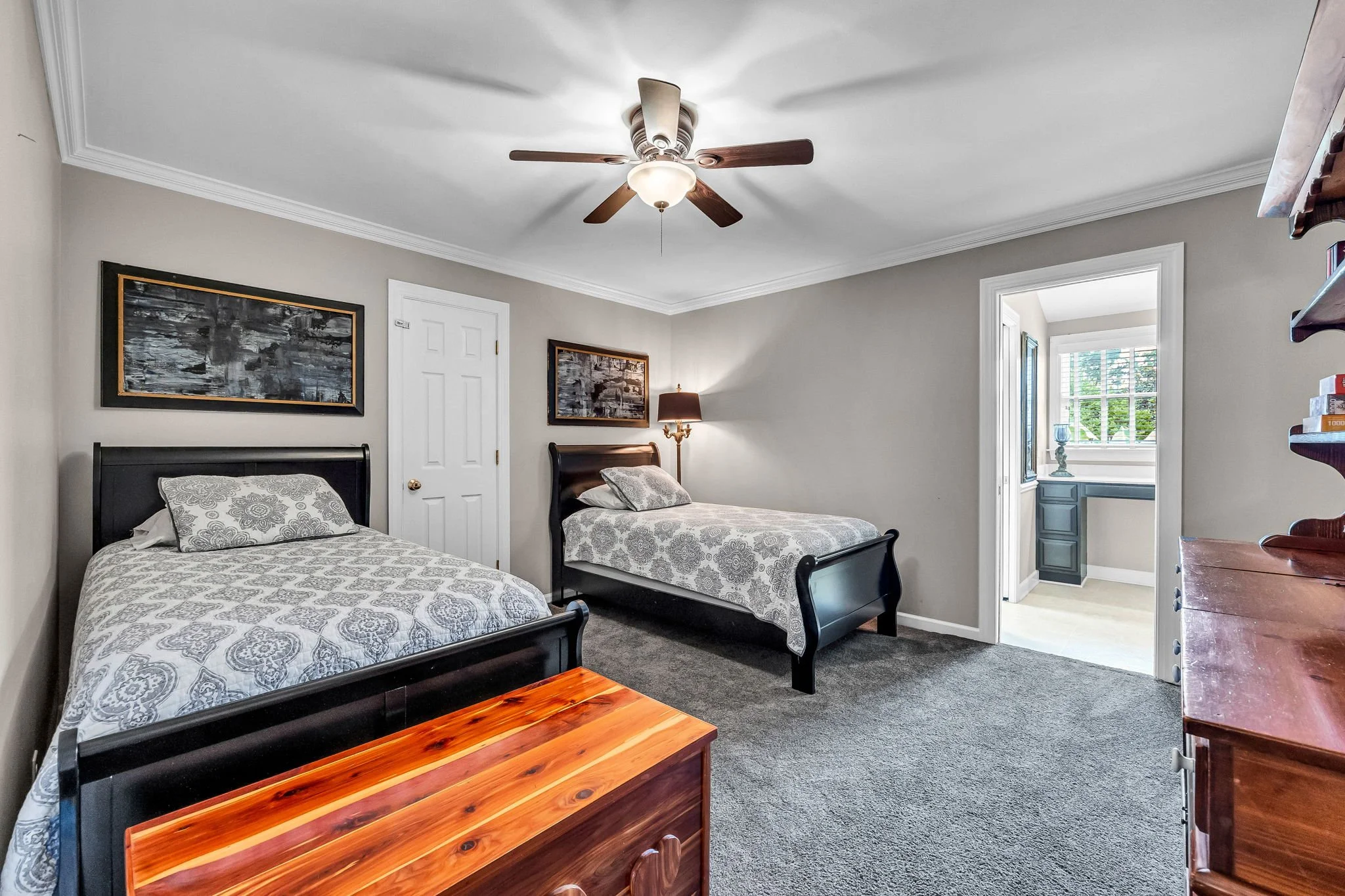 A bedroom with two twin beds with patterned bedspreads, a wooden dresser, a wall-mounted shelf, framed artwork, a ceiling fan with light, a standing lamp, and an open doorway leading to a room with a window.