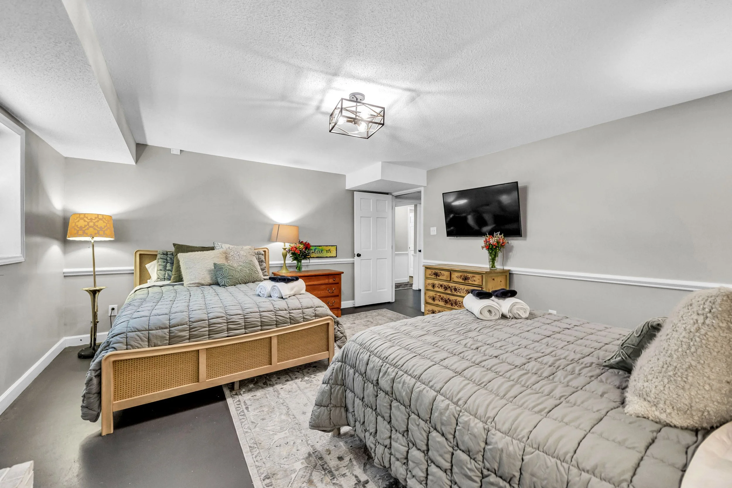 Two queen-sized beds with gray quilted bedding and pillows, each with rolled towels on top, in a bedroom with gray walls, a flat-screen TV on the wall, a wooden dresser, side table with flowers, and a ceiling light fixture.