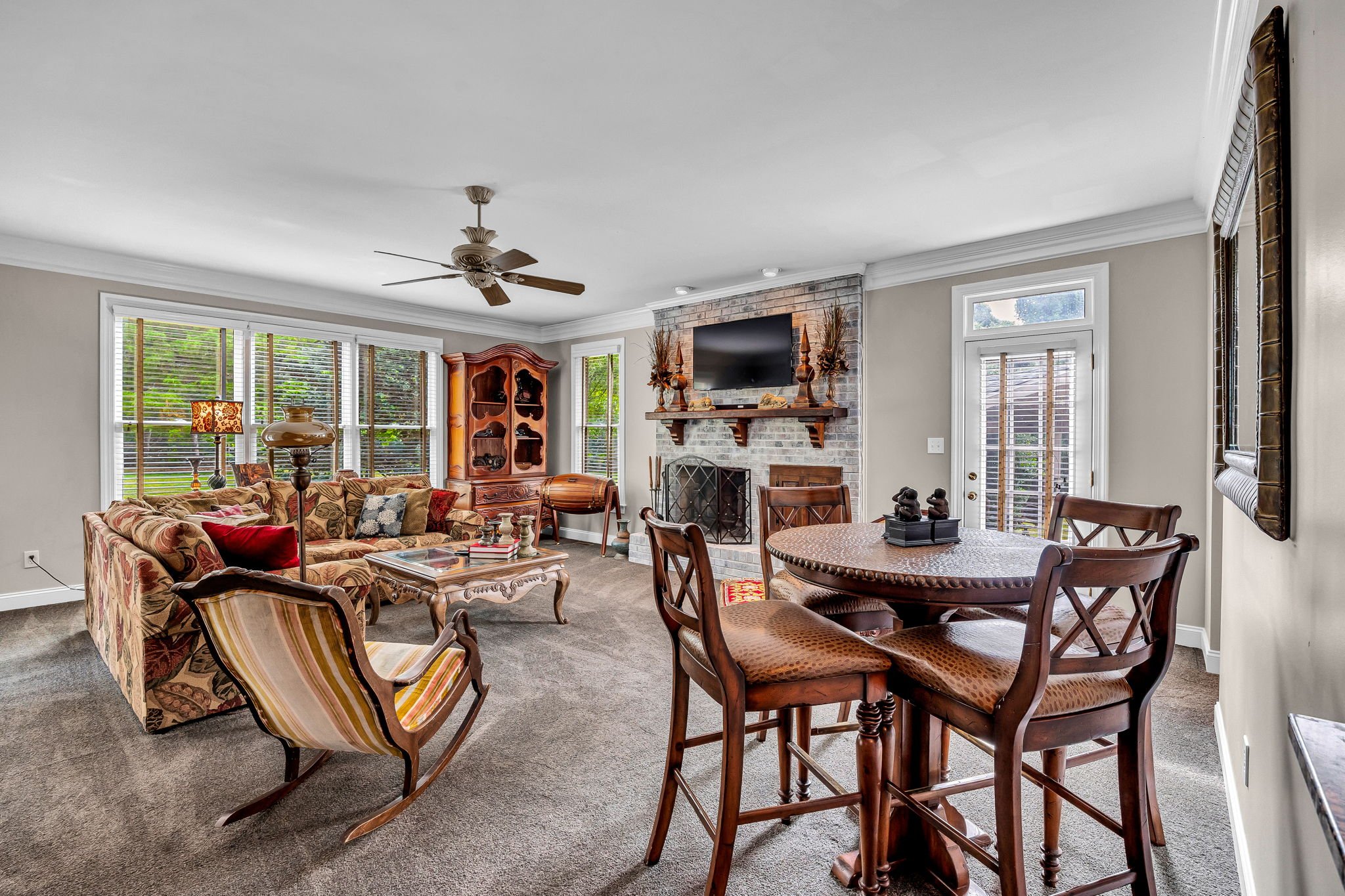 Living room with beige walls, large window with blinds, a ceiling fan, a brick fireplace with a TV on top, a floral patterned sofa, wooden chairs and a round dining table, decorative items, and a mirror on the wall.