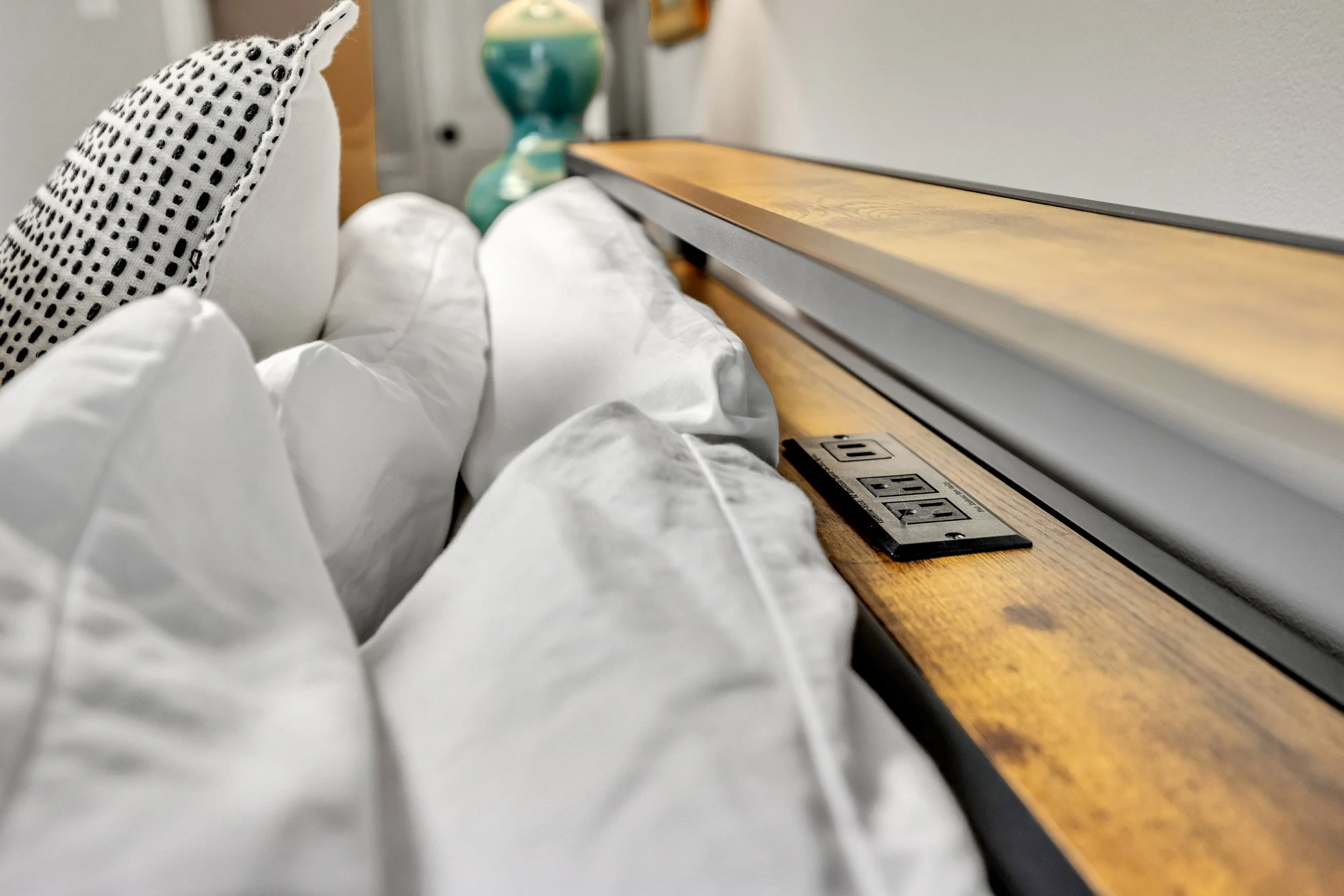 View of a hospital bed with white sheets and black, white, and gray polka-dotted pillow. The bed is positioned next to a wooden bedside table with electrical outlets and a black cable on the surface. In the background, there is a colorful ceramic vase and a white wall.
