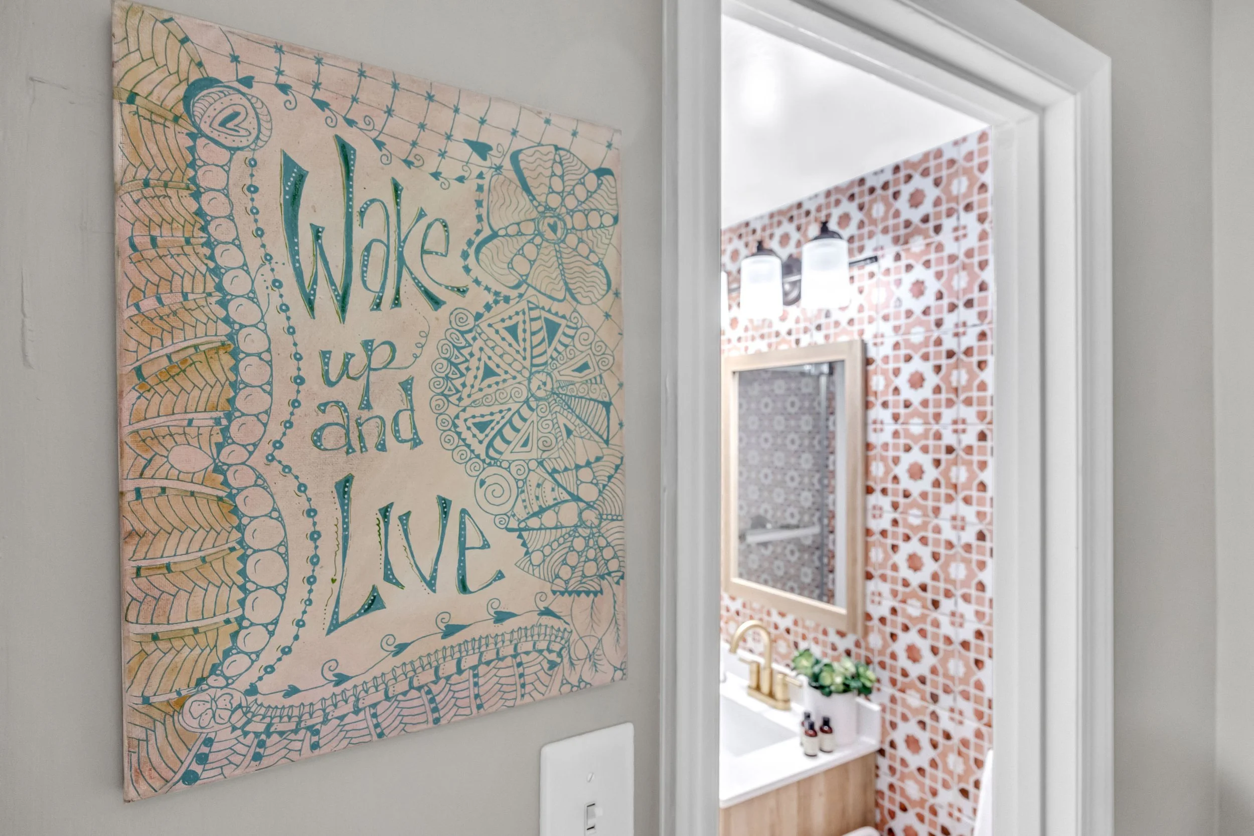 A decorative wall hanging with a floral and abstract design, and colorful text that says 'Wake up and live,' is displayed on a light-colored wall. A doorway leads to a bathroom with orange and white patterned tiles, a small window, a plant, and a countertop with toiletries.