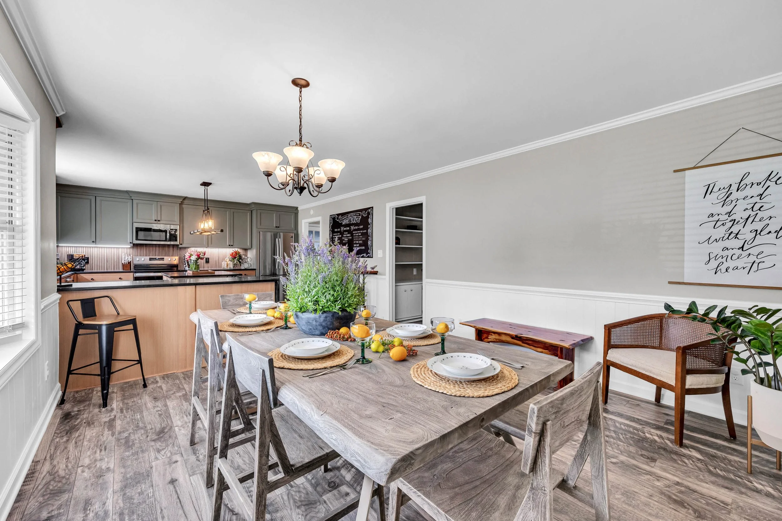 A dining area with a large wooden table set for a meal, surrounded by mixed chairs, in a room with a chandelier and natural light from a window, with a kitchen in the background featuring gray cabinets and stainless steel appliances.