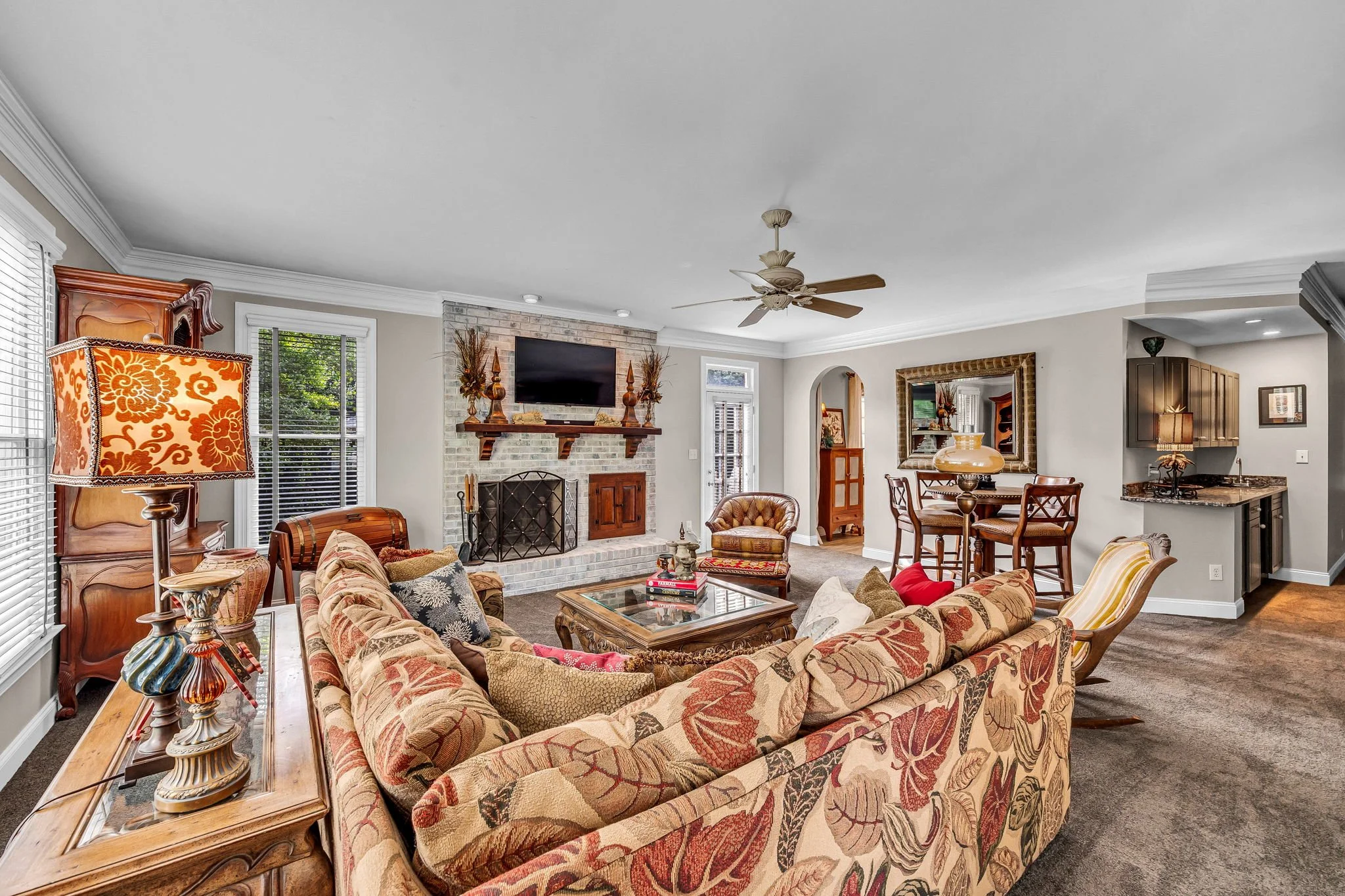 Spacious living room with floral-patterned furniture, a brick fireplace, a wall-mounted TV, and large windows with white blinds, decorated with lamps, vases, and wooden furniture.