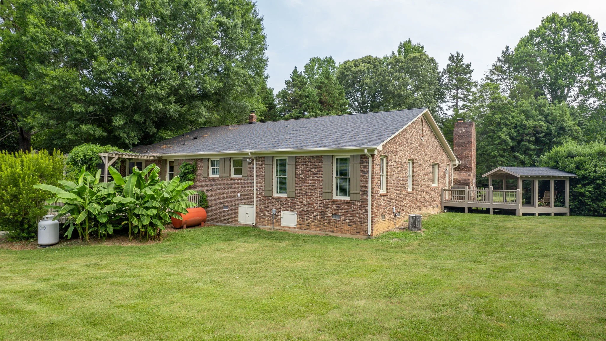 Backyard view of a brick house with a chimney, surrounded by lush green trees and grass, with a small deck and garden area.