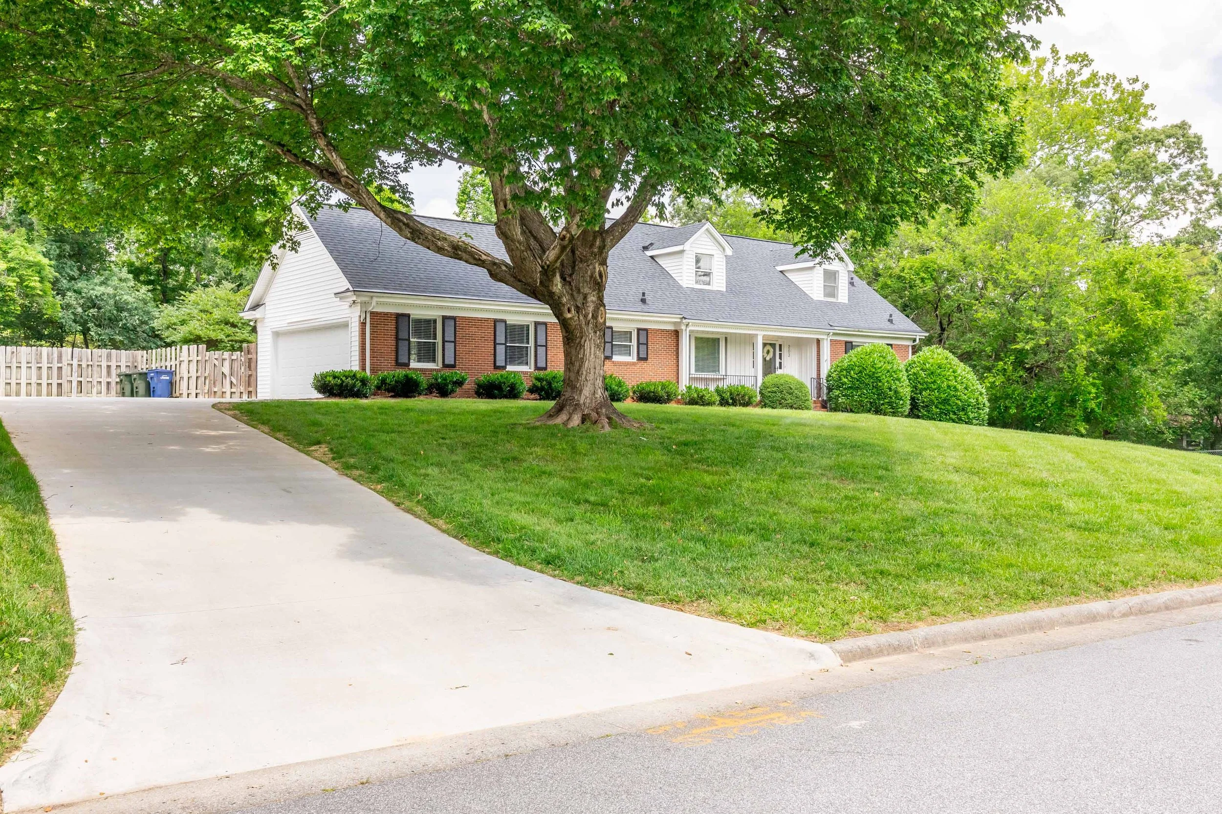 A suburban house with white siding and a brick facade, surrounded by green shrubs and a large tree in the front yard with a sidewalk and street in the foreground.