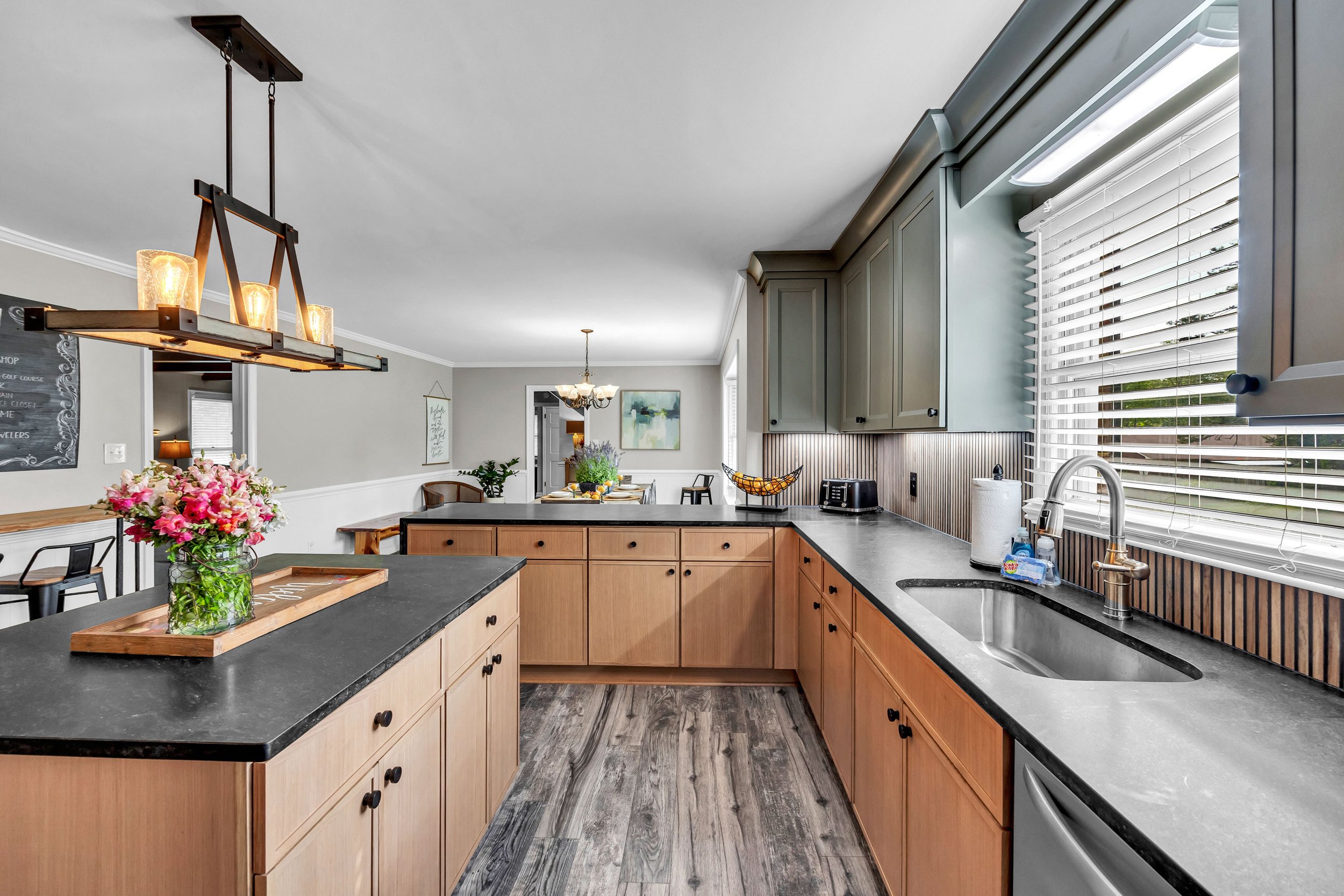 Kitchen with wooden cabinets, black countertops, stainless steel sink, and large window with blinds; dining area with chandelier and floral centerpiece.