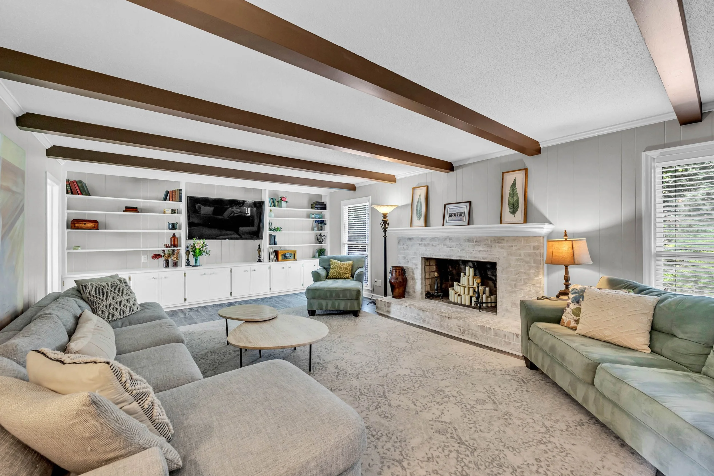 Living room with white walls, wooden ceiling beams, a fireplace with candles, shelf with books and decorations, green and beige sofas, a round coffee table, a lamp, and framed artwork.