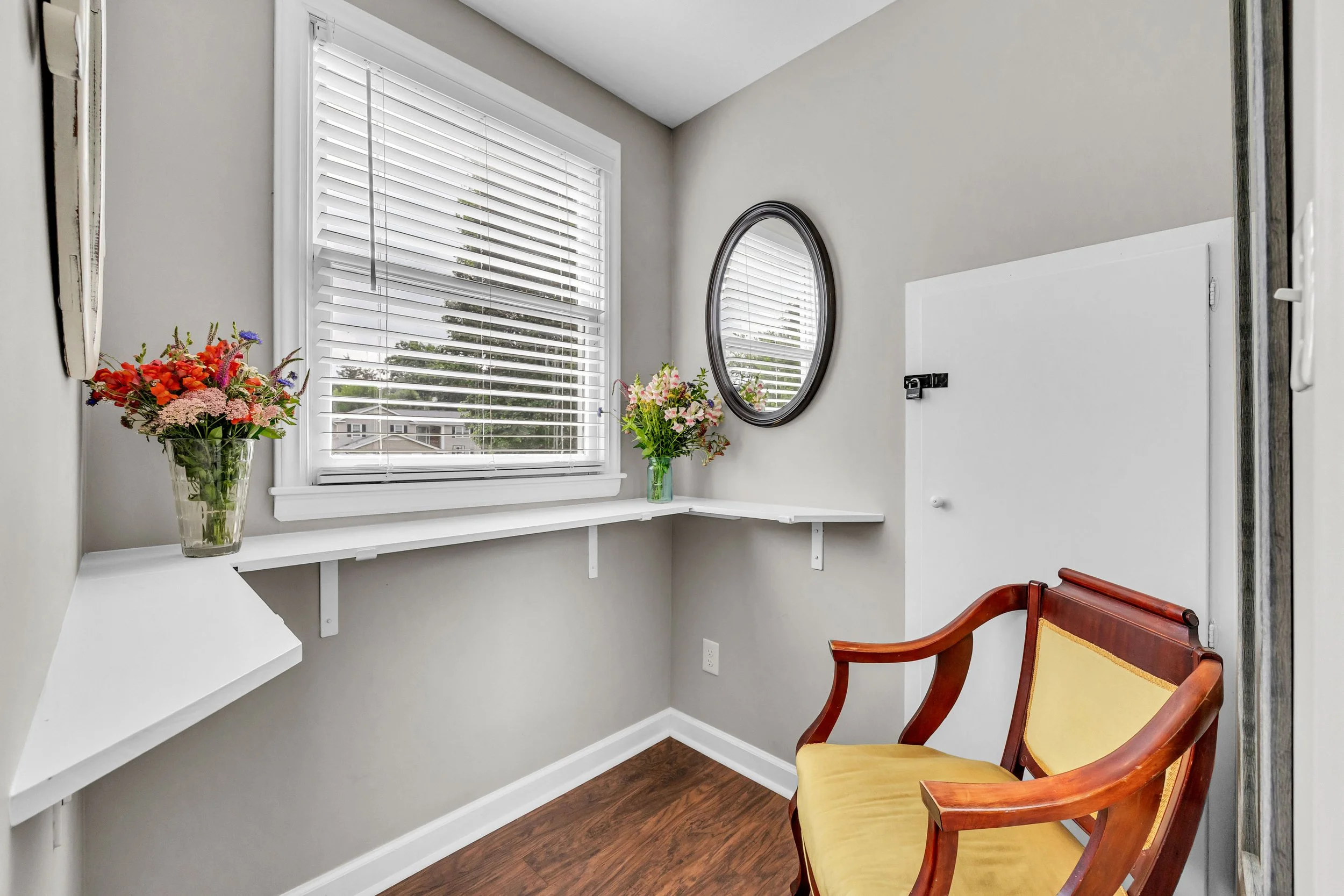 A small corner in a room with a window with blinds, two flower vases with colorful flowers on a white shelf, an oval mirror on the wall, a partial wooden chair with yellow upholstery, and a white door.