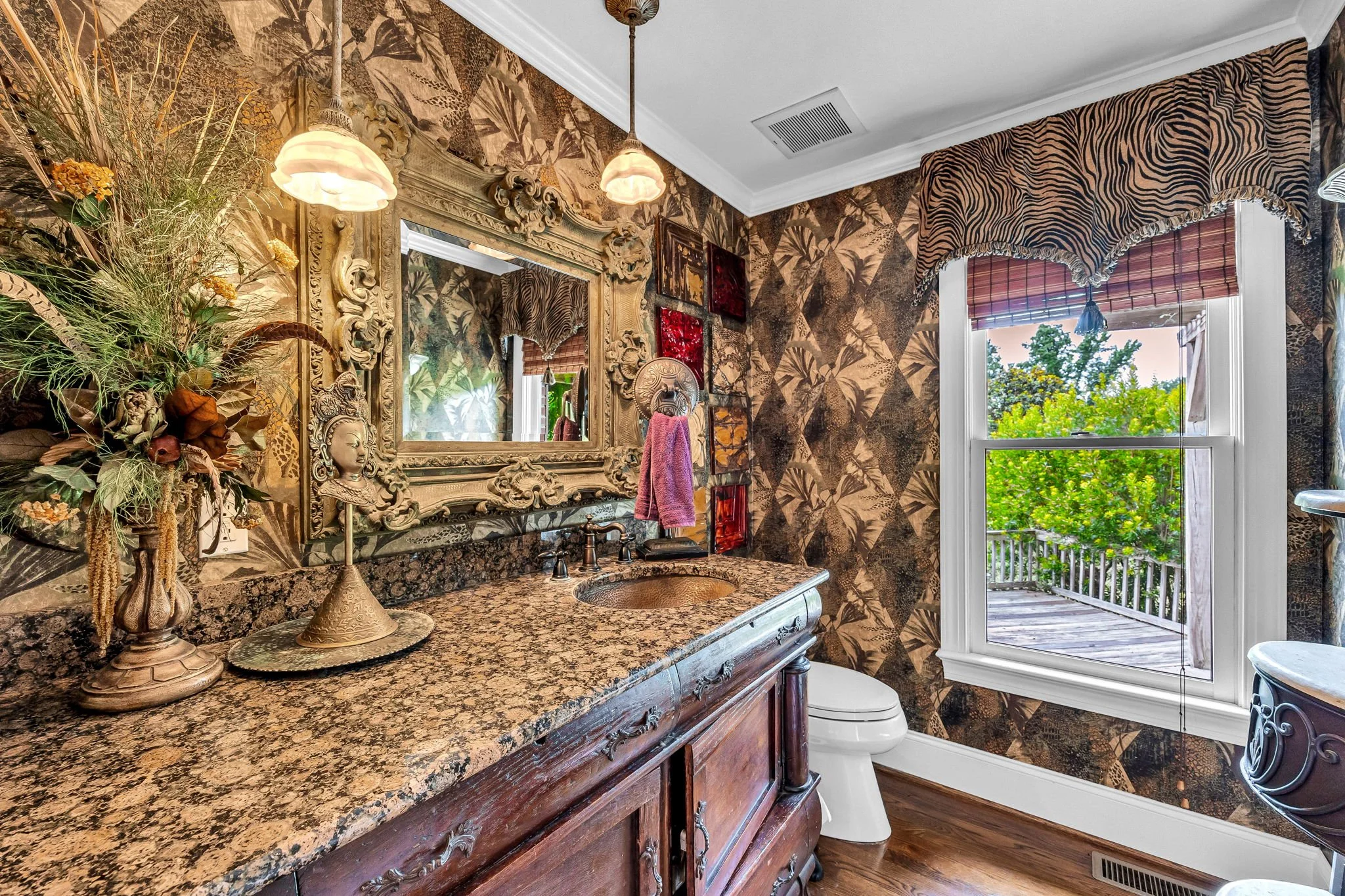 A bathroom with ornate decor, including a large gilded mirror, vintage-style vanity with a patterned granite countertop, and decorative wall art. There is a window with a zebra-print valance and a view of a balcony with green trees outside.