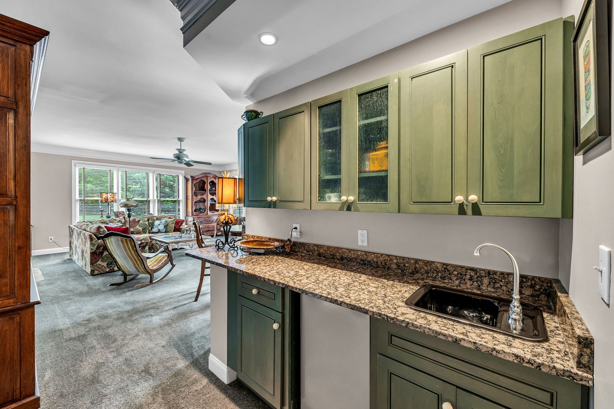 View of an open-concept living room and kitchen area with green cabinets, granite countertops, a black sink, and a living area with a sofa, chairs, and large windows that let in natural light.