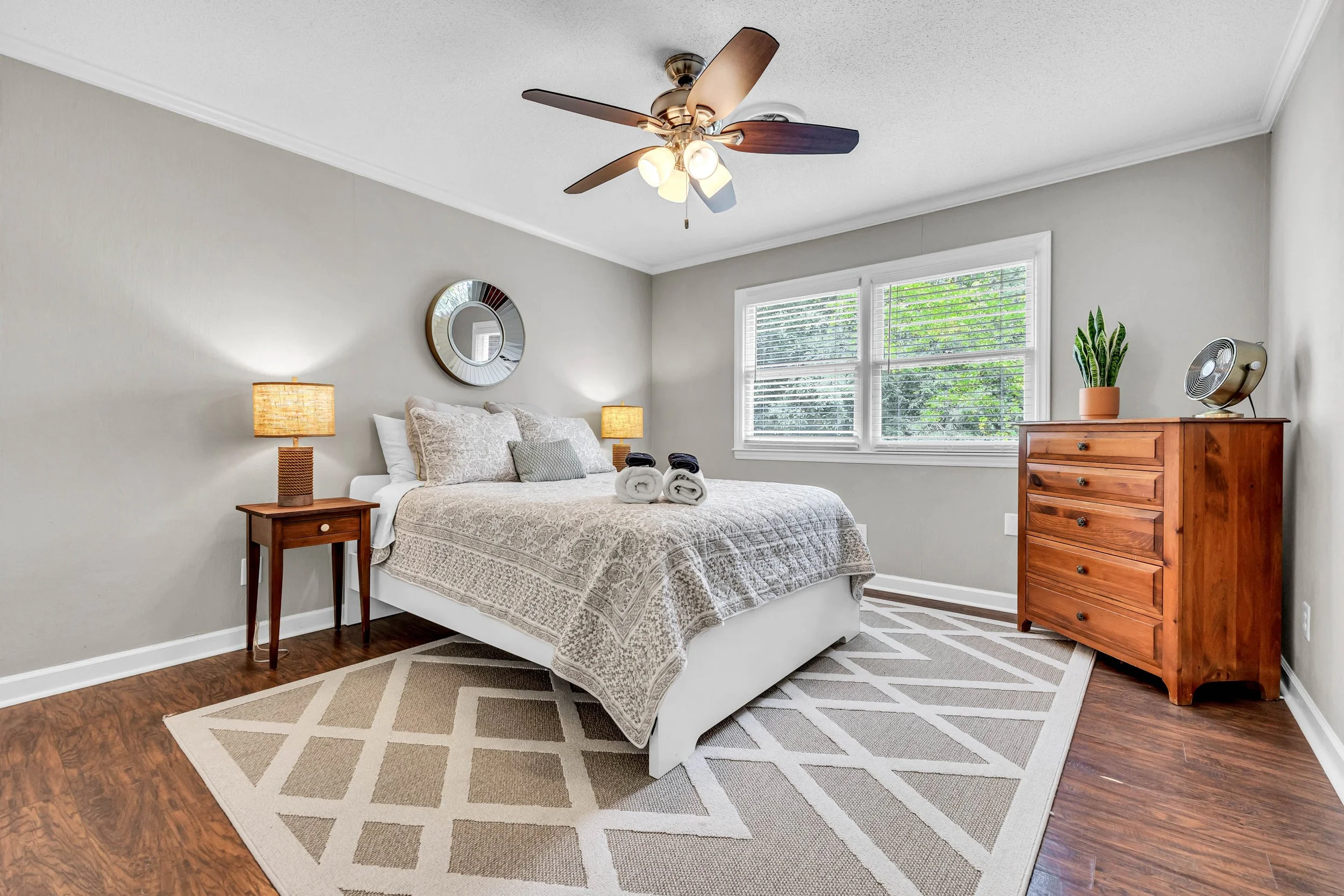 Bedroom with bed, nightstand, lamps, dresser, window, ceiling fan, mirror, potted plant, and a patterned rug.