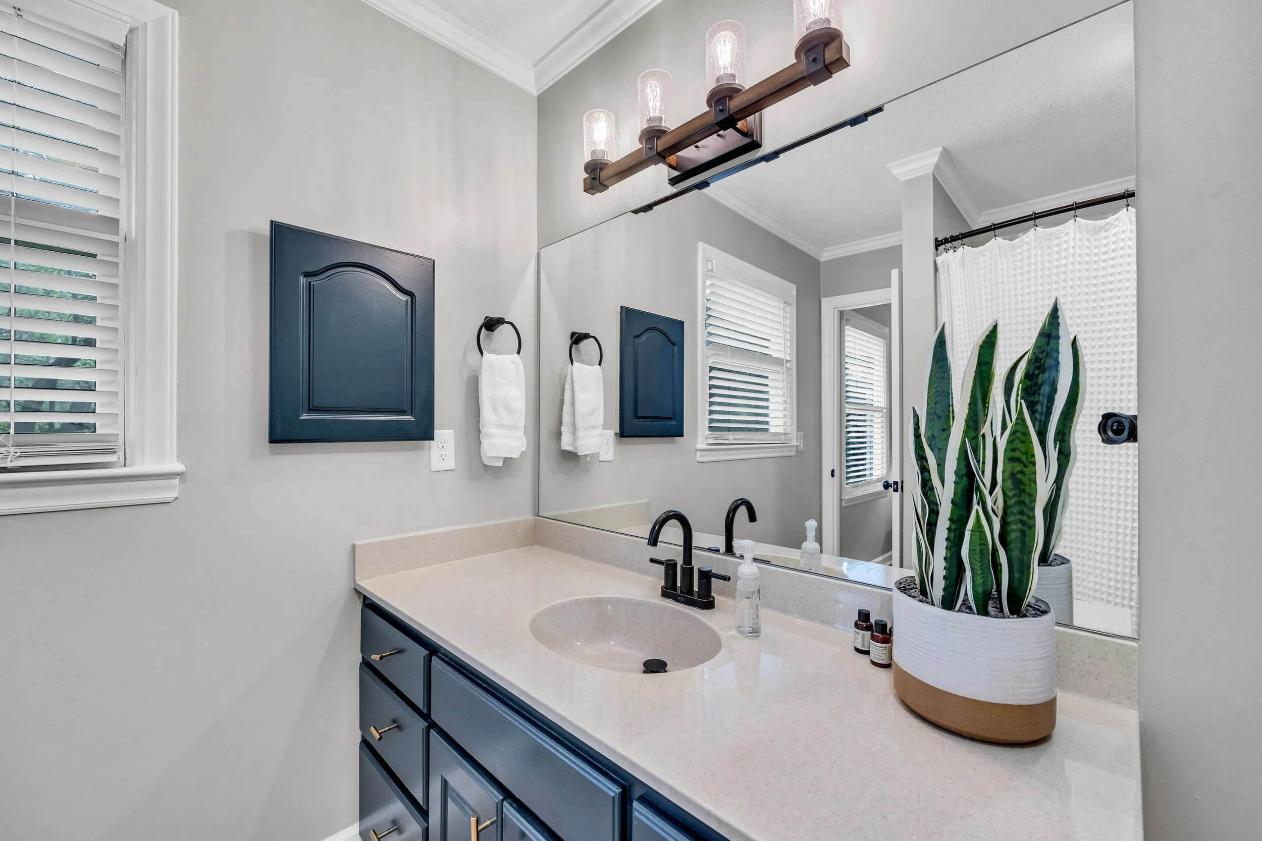 A bathroom with a large mirror above a beige countertop sink. There are two black faucets, soap dispensers, and small bottles on the counter. To the right, a tall potted snake plant is visible. The cabinet below the sink is painted navy blue, with gold handles. On the wall behind the counter, two blue cabinets are mounted, and a towel ring with white towels is visible. There are windows with white shutters and a door with a curtained window in the background. A wooden light fixture with four glass bulbs is mounted above the mirror.