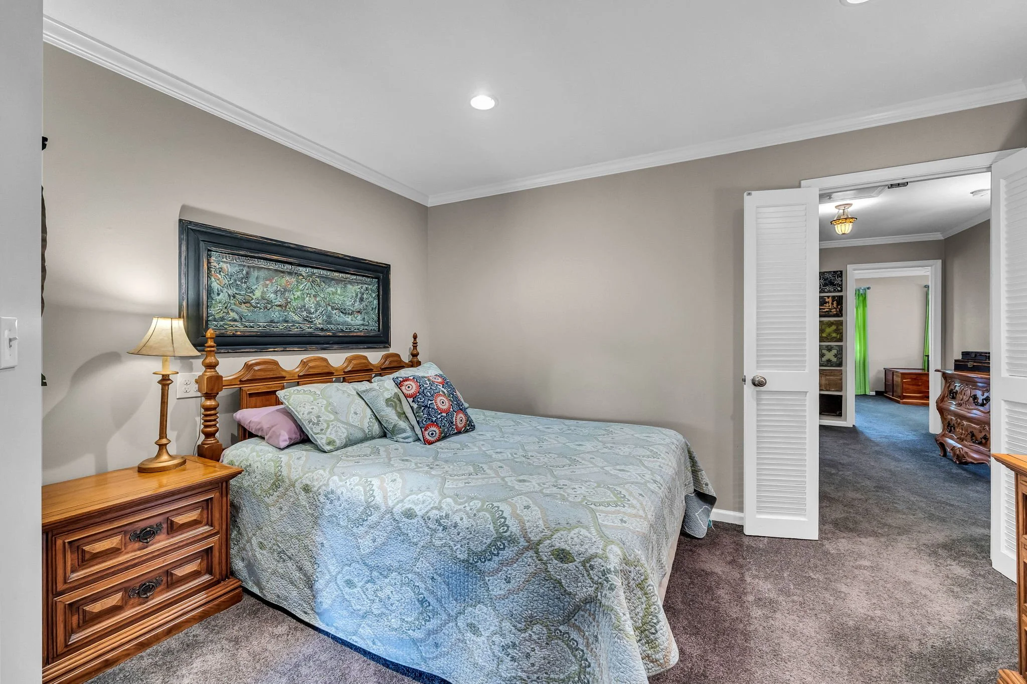 A cozy bedroom with a wooden bed frame, multicolored pillows, and a quilted bedspread. There is a nightstand with a lamp, a large framed picture above the bed, and an open closet with louvered doors leading to another room with green curtains and additional furniture.