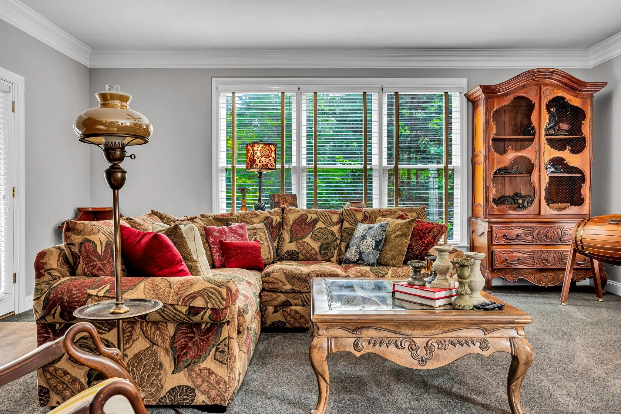 Living room with a floral-patterned sofa, wooden coffee table, antique china cabinet, and windows with blinds showing greenery outside.