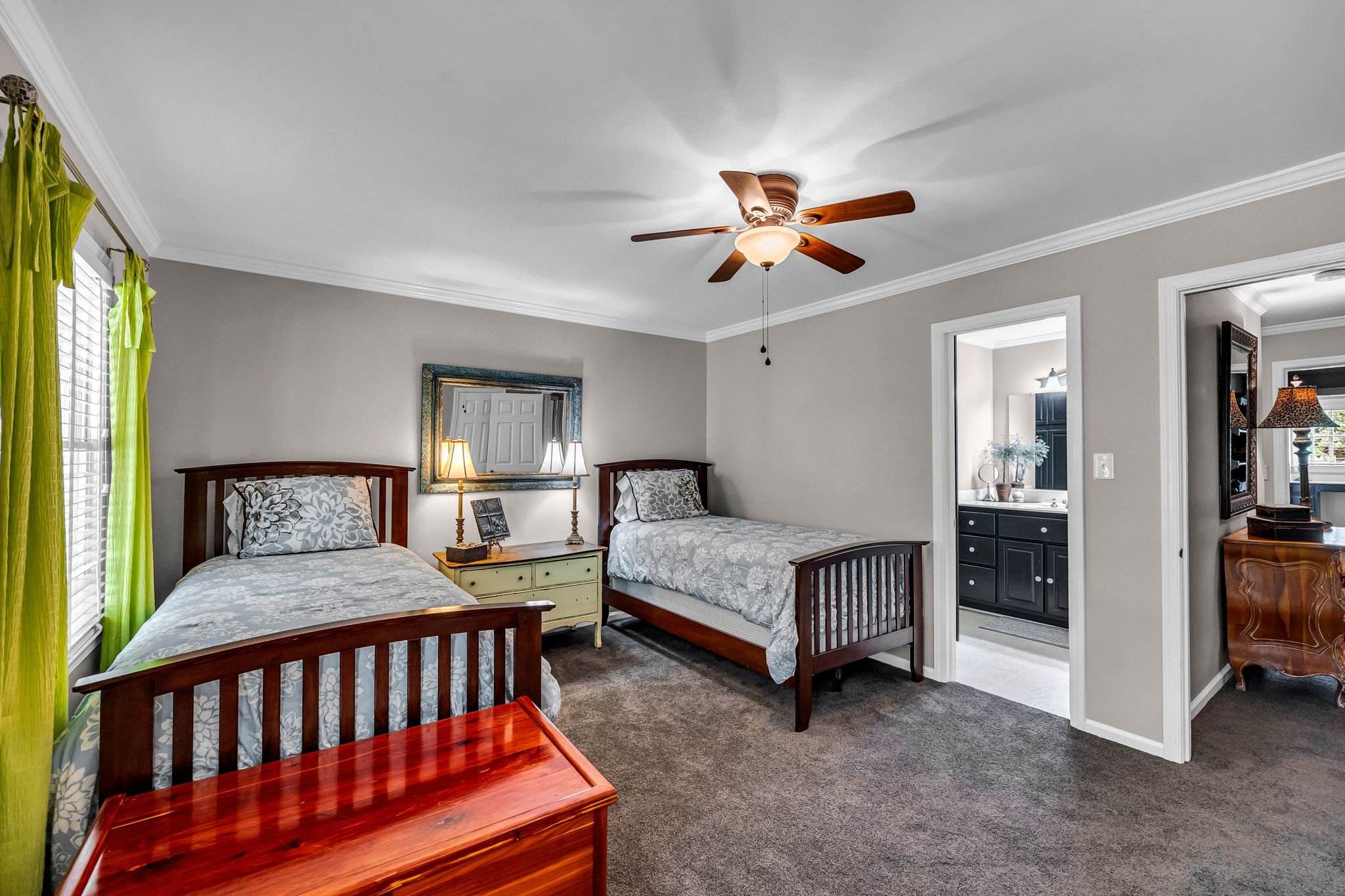A bedroom with two wooden beds, a yellow nightstand with lamps, a mirror, a window with green curtains, a ceiling fan, and a doorway leading to a bathroom with black cabinets and a white sink.