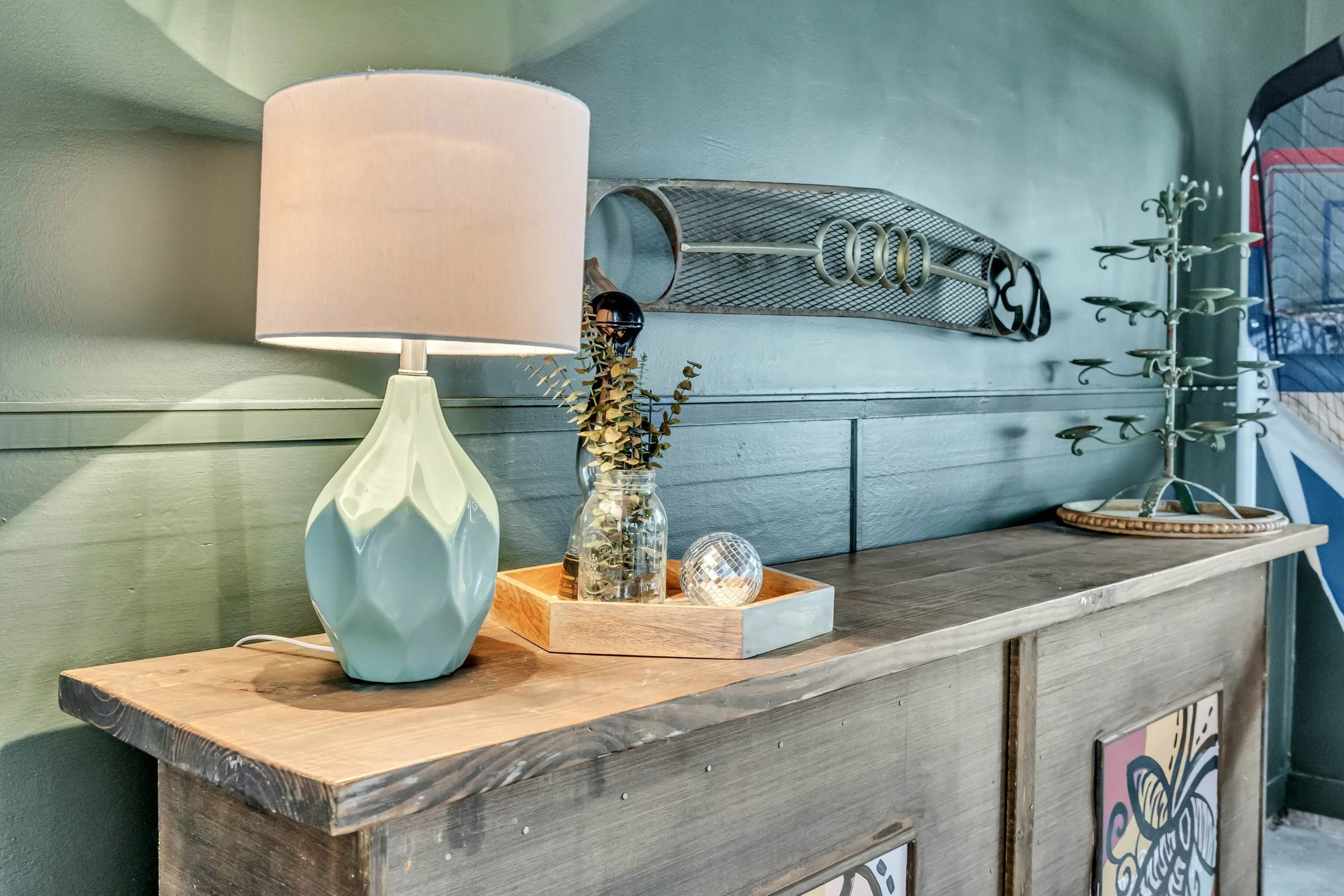 Wooden side table with a ceramic lamp, a small plant in a glass jar, a wooden tray, and decorative objects, against a green wall with metal wall art.
