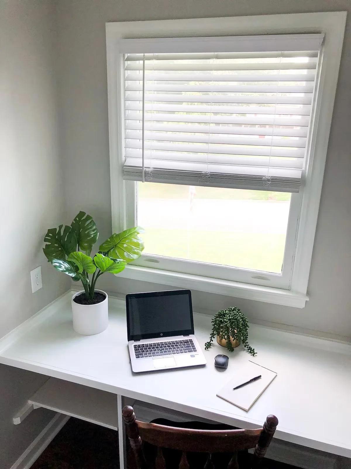 Home office workspace with a white desk, a laptop, a notepad with a pen, a computer mouse, a potted plant, a large leafy houseplant, and a window with white blinds.