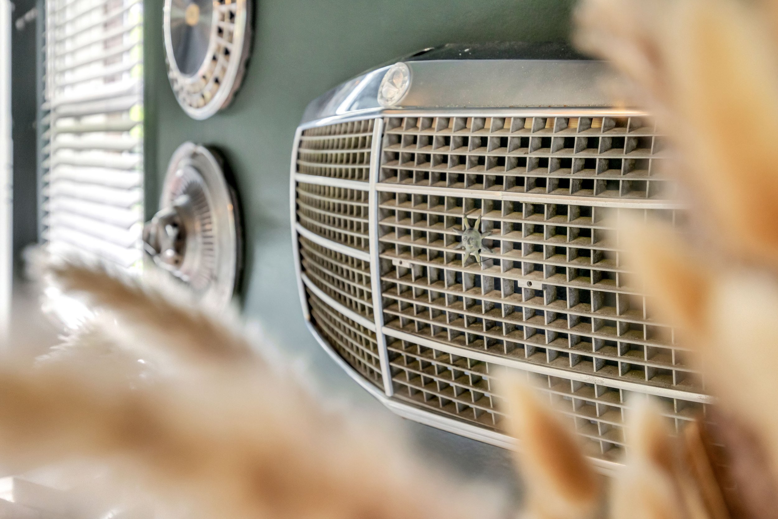 Close-up of the front grille of a vintage car, with a small emblem in the center, seen through a blurred foreground.