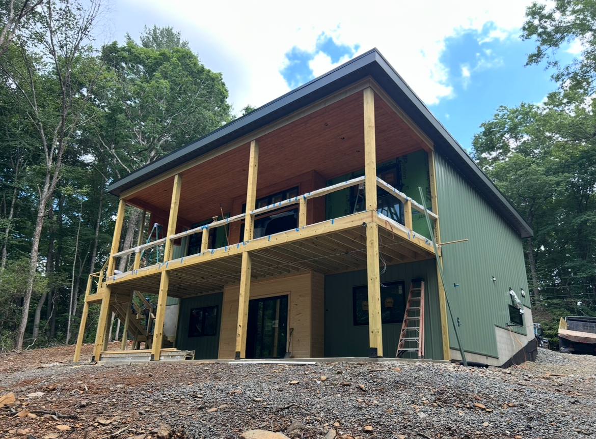 A two-story house under construction with a green exterior, wooden framing, and a balcony on the upper level. The house is in a wooded area with concrete stairs leading to the entrance.