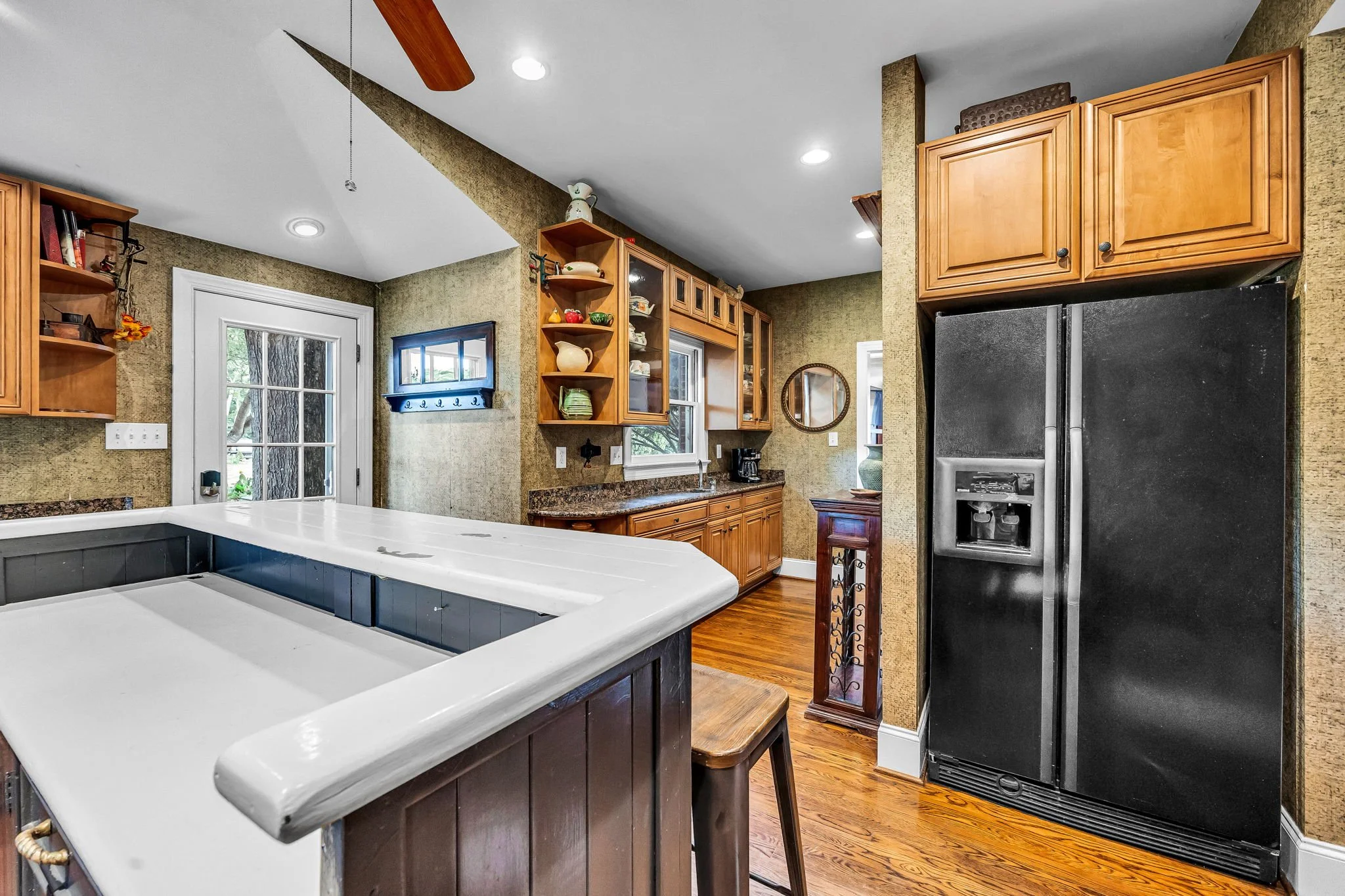 A kitchen with wooden cabinets, a black refrigerator, a white countertop, and hardwood floors. There are ceiling lights, a ceiling fan, and a small window with greenery outside.