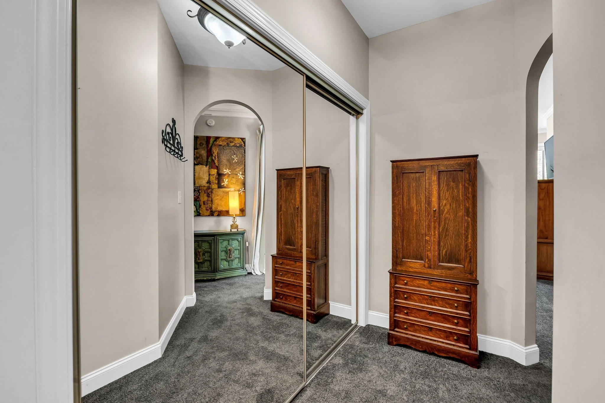 A room with a large mirror closet, wooden furniture, beige walls, and a carpeted floor. There is a glimpse into another room with a wall art and a lamp.