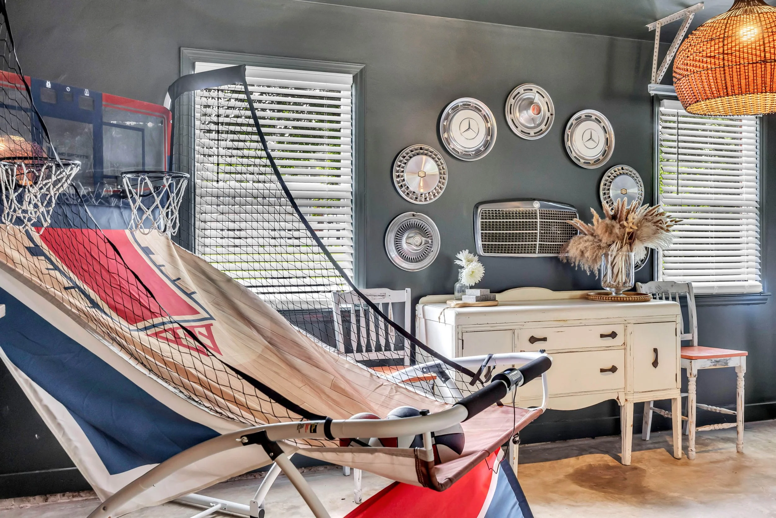 Indoor space with a blue and red basketball game setup, a white distressed cabinet, decorative wall mirrors, a table with a vase of dried flowers, and a white chair, with window blinds and a wicker lampshade in the background.