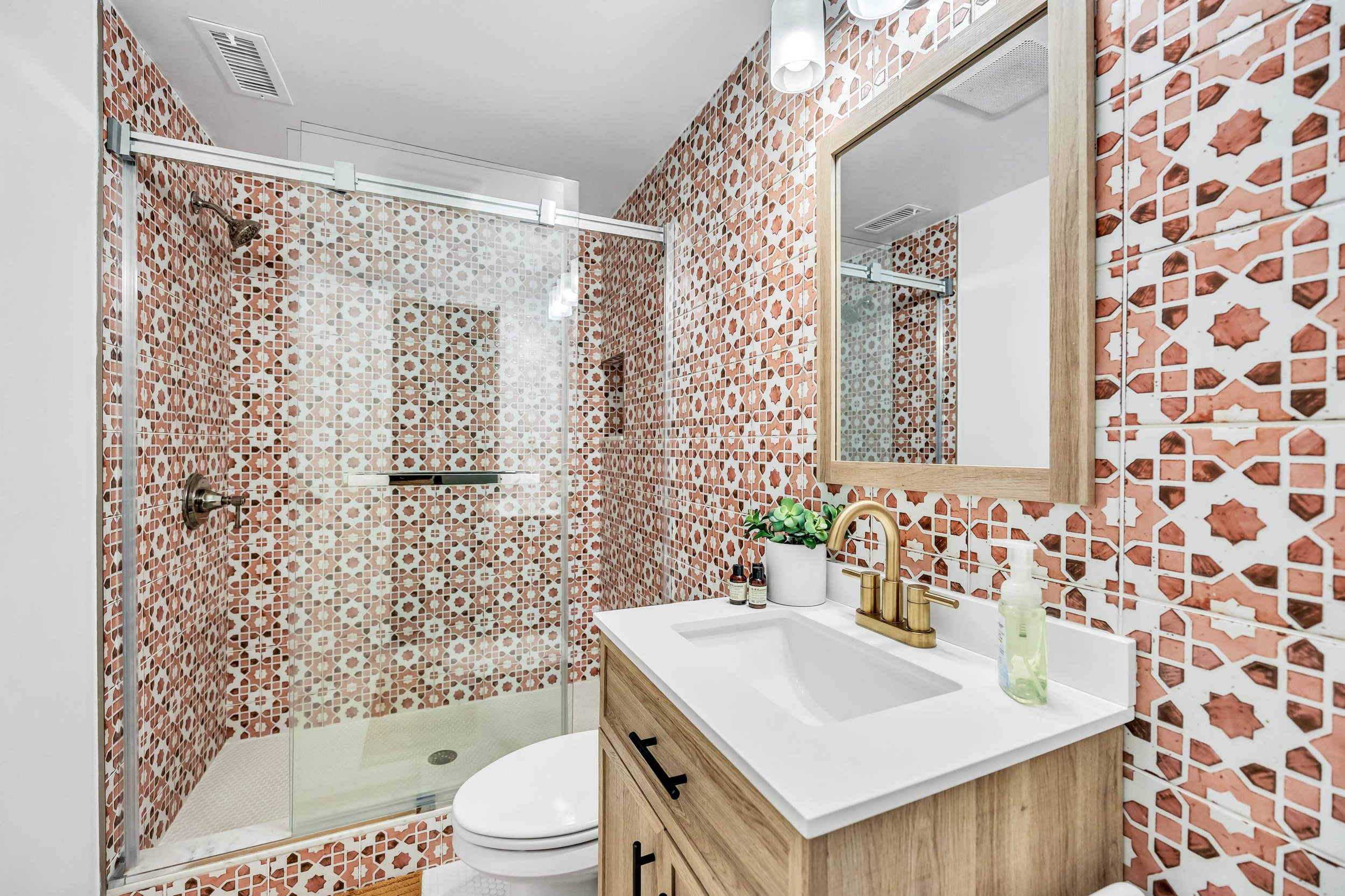 Bathroom with red patterned tile walls, glass shower enclosure, white sink with gold faucet, large mirror with wooden frame, and small potted plant on the sink counter.