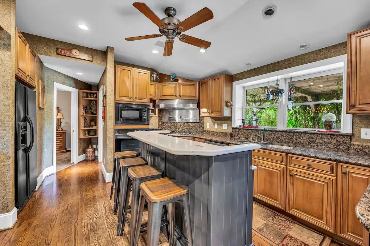 Kitchen with wooden cabinets, granite countertops, stainless steel appliances, a center island with four wooden seats, hardwood floors, and a window overlooking greenery.