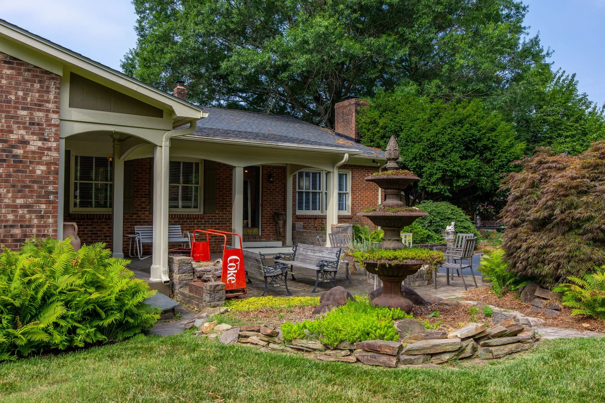 A backyard patio area with a stone fountain, multiple chairs, and lush green plants, with a brick house and large trees in the background.